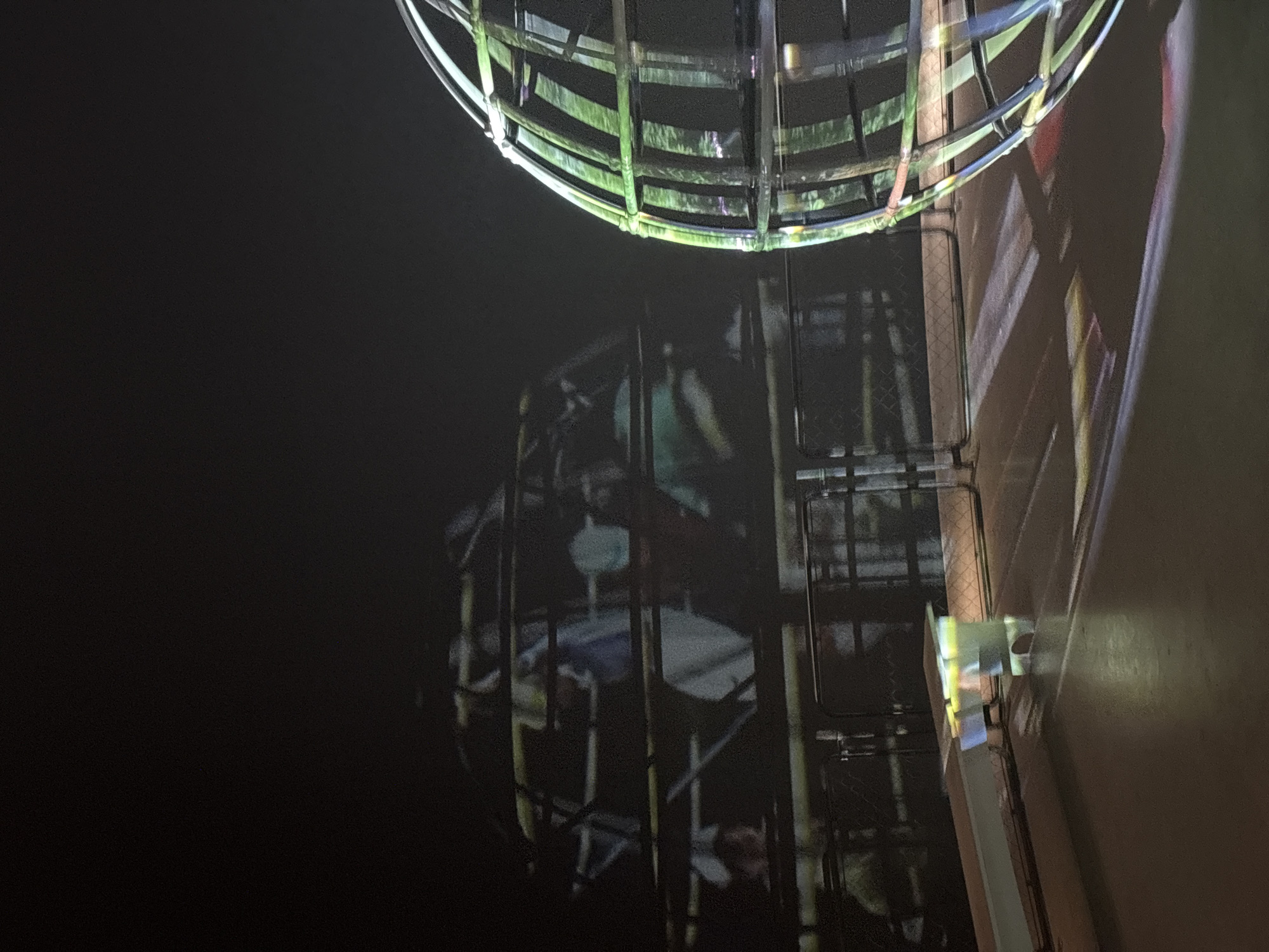 The image captures a night scene, likely an outdoor art installation or light show. Dominating the upper part of the frame is a large, spherical, cage-like structure, brightly illuminated with a greenish light. Its metallic or plastic bars form a geodesic-like pattern.
Below this structure, on a dark, possibly paved or wall surface, is a large projection or shadow. This projection depicts what appears to be human figures or silhouettes enclosed within a similar, dark, cage-like framework. The figures are indistinct but suggest people standing or moving within the projected structure.
To the right, a muted, reddish-brown wall or building facade runs vertically, partially illuminated by various light sources, creating abstract patterns of light and shadow in shades of yellow, blue, and white. A small, light-colored fixture is visible on this wall, seemingly emitting some of the light. Further down and to the left of the wall, there's a glimpse of what looks like a fence or gate with a grid pattern.
The overall impression is one of a dramatic interplay of light and shadow, creating a sense of depth and artistic intrigue in a dimly lit environment. The time of day is clearly night. No visible text is present.