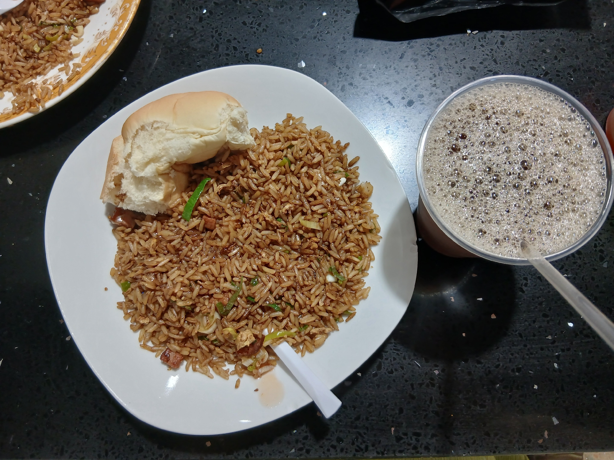 The image displays an overhead view of a meal set on a dark, speckled countertop, likely in Juan Griego, Venezuela. The central focus is a white, square-shaped plate laden with fried rice. The rice is a warm brown hue, indicating the use of soy sauce, and is mixed with finely chopped green onions or other vegetables and small pieces of meat. A soft, light-colored bread roll or bun rests atop the rice. A small, folded white plastic item, possibly a condiment packet or wrapped utensil, lies on the plate near a small reddish liquid mark.
To the right of the main plate, a clear plastic cup holds a dark, bubbly beverage, likely a soda or coffee drink, with a straw inserted. A second, partially visible plate with a gold and white patterned rim is located in the upper left corner, also containing fried rice. The countertop appears to be made of a stone material like granite or quartz. The scene depicts a ready-to-eat meal, with no individuals visible. There is no discernible text in the image.
