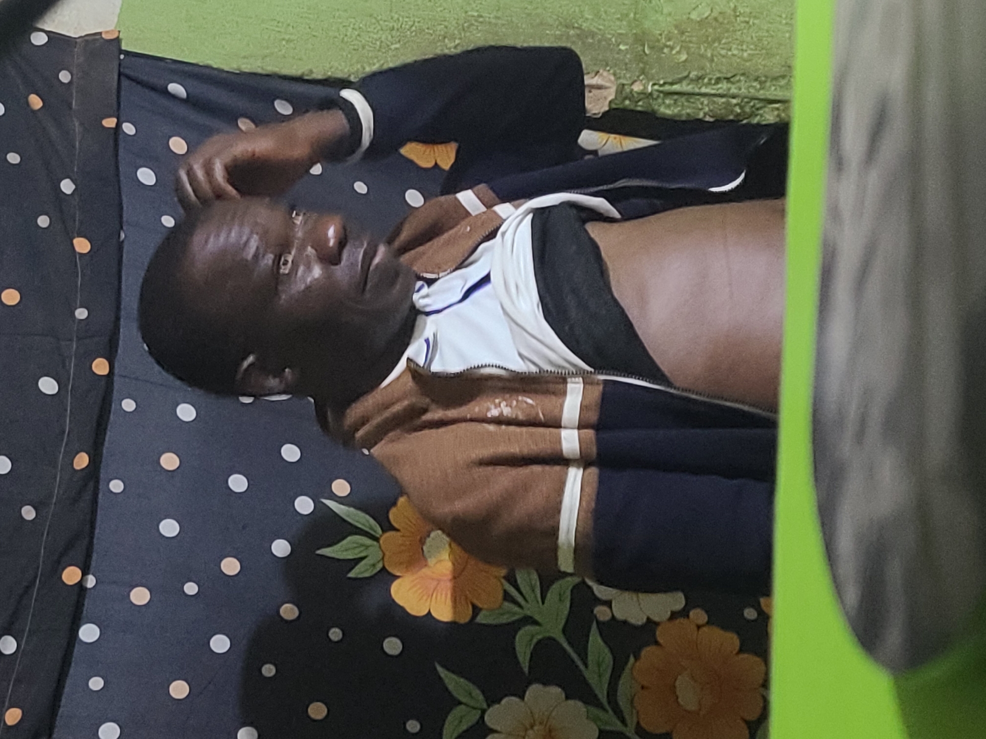 An adult Black male is lying on a bed with a dark floral and polka dot patterned bedsheet, wearing a partially unzipped, brown and navy jacket over a white top. The scene appears to be indoors, with a green painted wall visible in the background. The location is stated to be Jos, Nigeria.