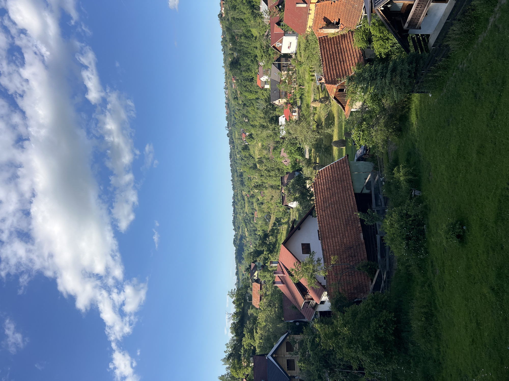 The image is a photograph taken from a high vantage point, looking down on a rural village nestled in a lush, green valley. The sky is a clear, bright blue with scattered white clouds. The village consists of numerous houses with reddish-brown tiled roofs, interspersed with abundant trees and vegetation. The houses vary in size and architectural style, with some appearing to be older, traditional homes and others more modern. Many of the houses are situated on sloping terrain, suggesting a hilly or mountainous region.
The scene appears peaceful and idyllic, indicative of a rural setting during daylight hours. The bright blue sky and scattered clouds suggest fair weather. The presence of hay bales in some of the grassy areas might suggest the time of year is late spring or summer, during or just after haymaking season. There is no visible text in the image. The overall impression is one of a serene, lived-in rural community surrounded by nature.