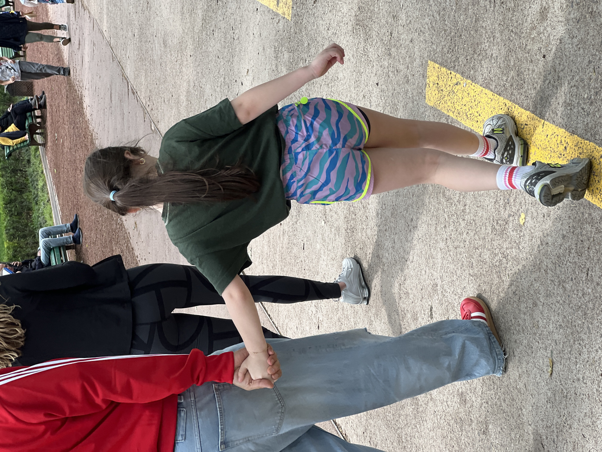 A girl in athletic shorts stands on a sidewalk with yellow stripes, next to a man in jeans and a red jacket, holding hands. Other people are sitting on benches and walking in the background. The location is possibly in Tbilisi, Georgia.