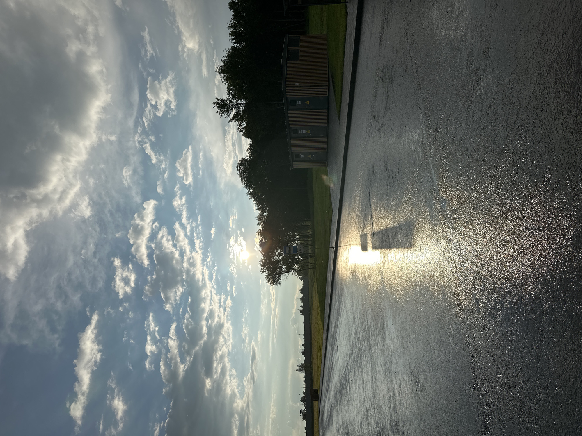 The image depicts an outdoor scene, likely at a park or recreational area. The perspective is a low-angle, sideways shot, with the sky and a paved surface dominating the frame.
The sky is filled with a mix of bright white and darker grey clouds, suggesting a partly cloudy day. The sun is visible, shining brightly and casting a strong glare on the wet pavement in the foreground. This glare creates a reflective sheen on the dark asphalt, highlighting its wetness and texture.
In the background, a line of trees forms a dense, dark silhouette against the sky. Partially visible amidst the trees and grass is a small, modern-looking building with a wooden exterior, featuring multiple doors and what appear to be warning symbols. There is also a signpost with text, though the text is too small to read. A shadow of an object, possibly a lamppost or a similar structure, is cast onto the wet pavement, further emphasizing the presence of the sun.
The wetness of the pavement and the bright sunlight suggest that it may have recently rained, or that the area is being watered. The overall mood is serene and slightly dramatic due to the strong contrast between light and shadow. The time of day appears to be late afternoon or early evening, given the angle of the sun and the quality of the light. No people are visible in the image.