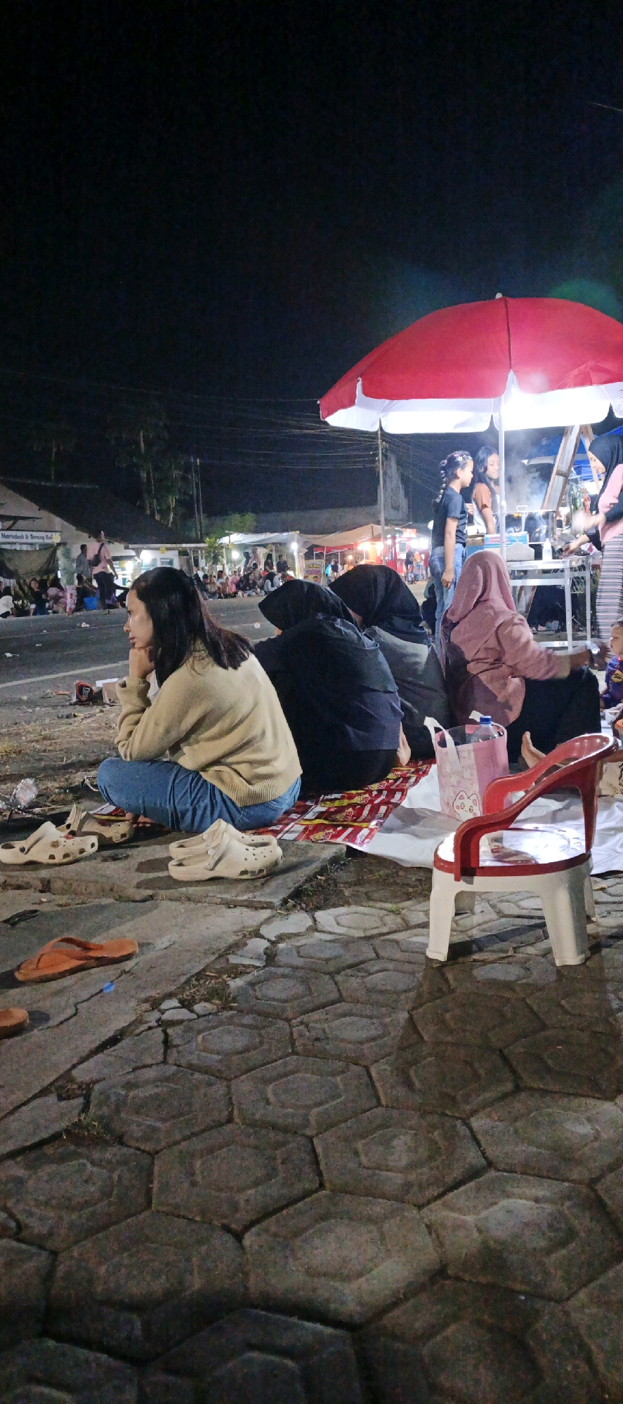 The image depicts a nighttime street scene in Kasiyan, Indonesia, with a focus on a group of people seated on the ground. A red and white umbrella, likely belonging to a food stall, is visible at the top right of the frame. In the foreground, a woman in a beige sweater and blue jeans is seated with her elbow on her knee and chin resting on her hand, looking to her left. To her right, two other individuals are seated, both wearing hijabs; one in black and the other in a mauve hijab. A white plastic chair with red accents sits empty in front of them. Scattered on the ground are several pairs of shoes, including white clogs and orange flip-flops. Behind the seated individuals, other people are visible, some sitting and some standing, in front of various stalls or shops that are illuminated by lights. A road is present in the middle ground, and beyond that, there are buildings and trees under a dark night sky. The ground is paved with hexagonal stones.