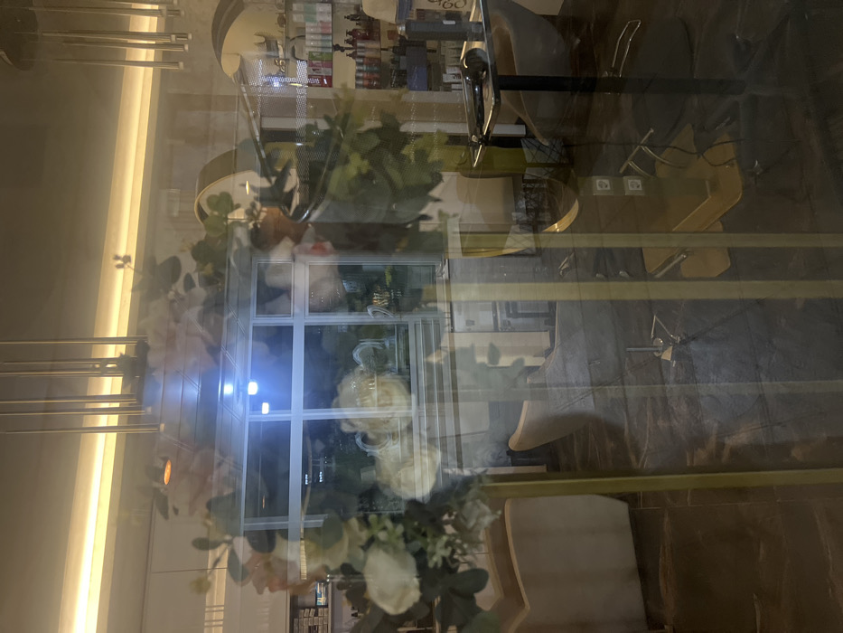 The image displays the interior of a hair salon or beauty parlor, viewed through a reflective glass partition in Smederevska Palanka Municipality, Serbia. The foreground is dominated by reflections on the glass, primarily featuring a decorative floral arch or frame composed of white and pink flowers with green foliage, supported by vertical gold-colored elements. Reflected in the upper center is a multi-paned window with bright blue illumination from beyond.
Within the salon, visible through the reflections, are various elements. On the right, shelves are stocked with numerous bottles of hair care products. A dark salon chair with a footrest and a small dark table holding a hairdryer and brush are present. A large, curved mirror with a gold frame is partially visible. On the left, light-colored upholstered chairs or seating are arranged on a dark floor. The ceiling features multiple hanging gold-colored cylindrical light fixtures and a continuous strip of warm yellow lighting along the perimeter. The environment appears modern and well-lit. No individuals are visible within the scene or its reflections.