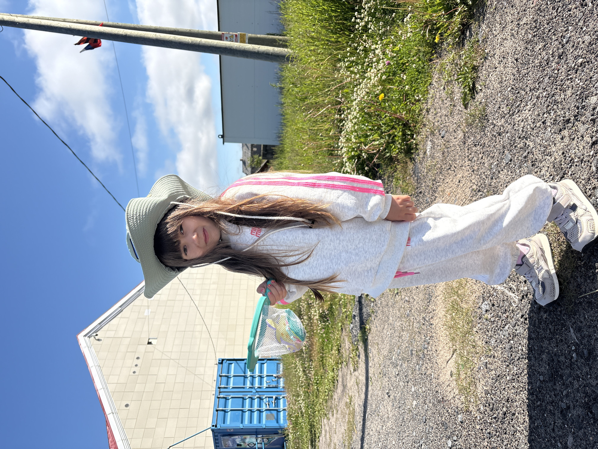 A young girl wearing a light blue wide-brimmed hat, a gray tracksuit with pink stripes, and gray sneakers stands in an outdoor setting. She holds a green-handled butterfly net filled with colorful objects. The ground is covered in gravel and sparse green grass with small white flowers. In the background, there is a large, light-colored building with a red roof and a blue shipping container. Further back, a tall wooden pole with two flags and a white building can be seen. The sky is blue with scattered white clouds. The scene appears to be in the city of Teriberka, Russia, given the context.