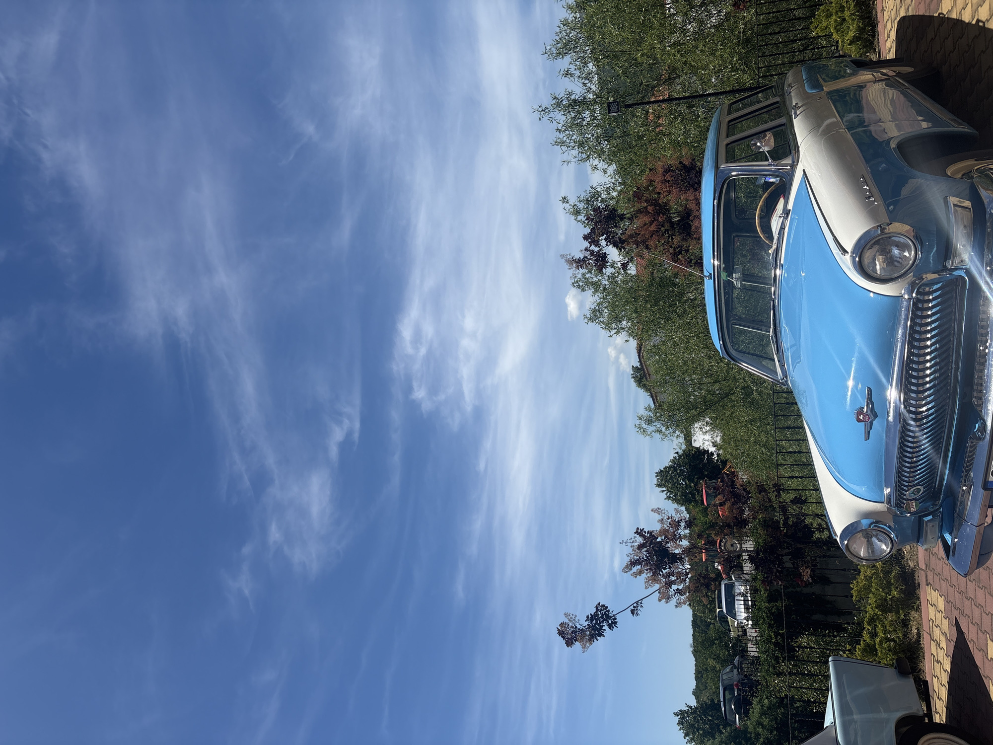 The image shows a blue and white vintage car parked in an outdoor location. The sky is predominantly blue with wispy clouds. The car is positioned near foliage, including trees and shrubbery, and part of a brick-paved area is visible. Other vehicles are visible in the background, parked behind a fence. The location is described as Unknown, Unknown.