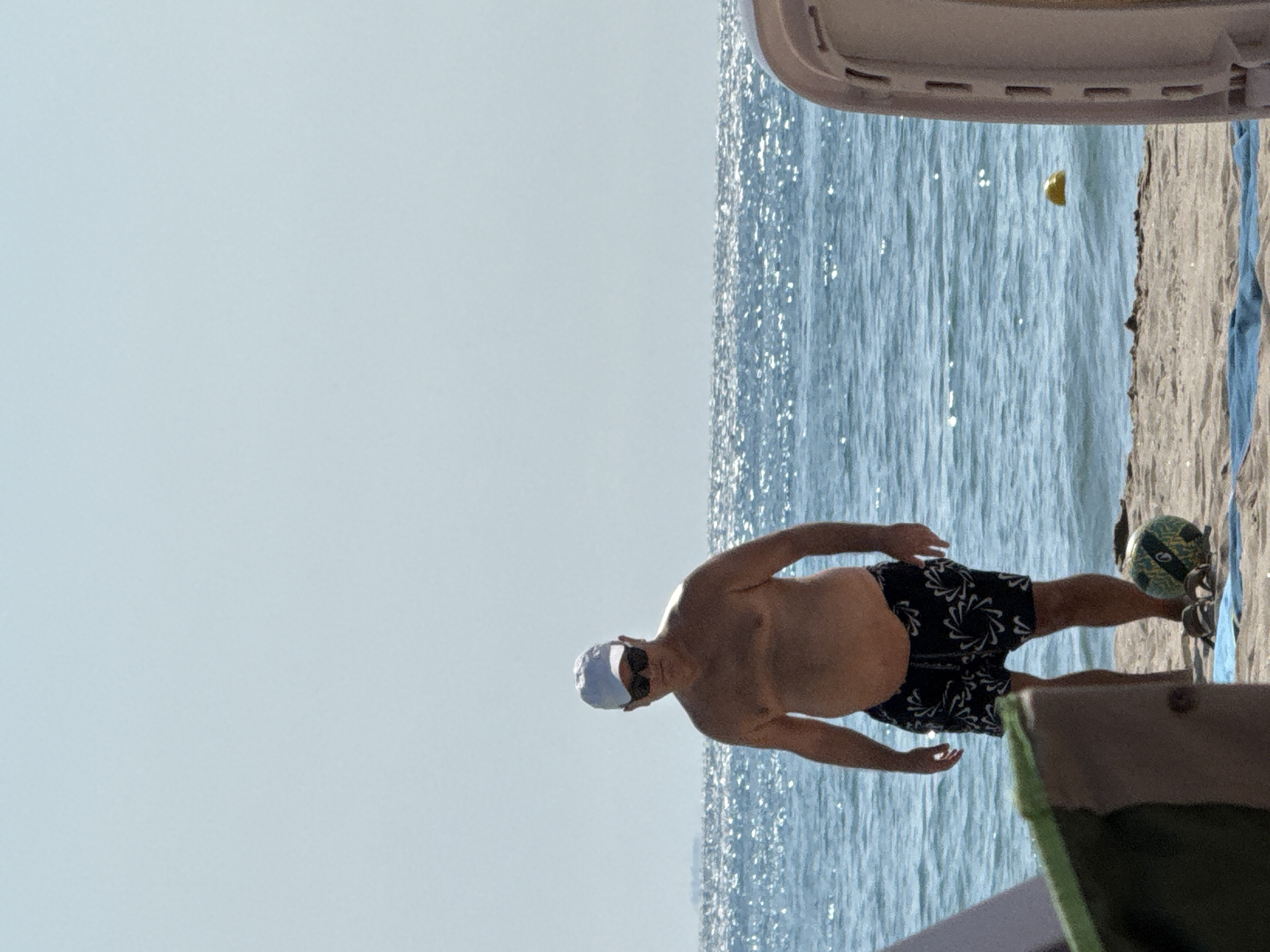 A shirtless, overweight man wearing a white baseball cap, sunglasses, and black and white patterned swim trunks stands on a sandy beach facing the camera. He is positioned in the lower-left quadrant of the frame. The man's hands are held loosely at his sides. To his right, at his feet, is a dark-colored ball with green accents and a pair of flip-flops on a blue towel. The ocean stretches behind him, with its surface sparkling under the sun. A yellow buoy is visible floating in the water to the right of the man. The sky above is a pale blue, nearly cloudless. Parts of beach chairs are visible in the foreground and on the right side of the image, partially obscuring the scene. The location is the coast of Obrochishte, Bulgaria.