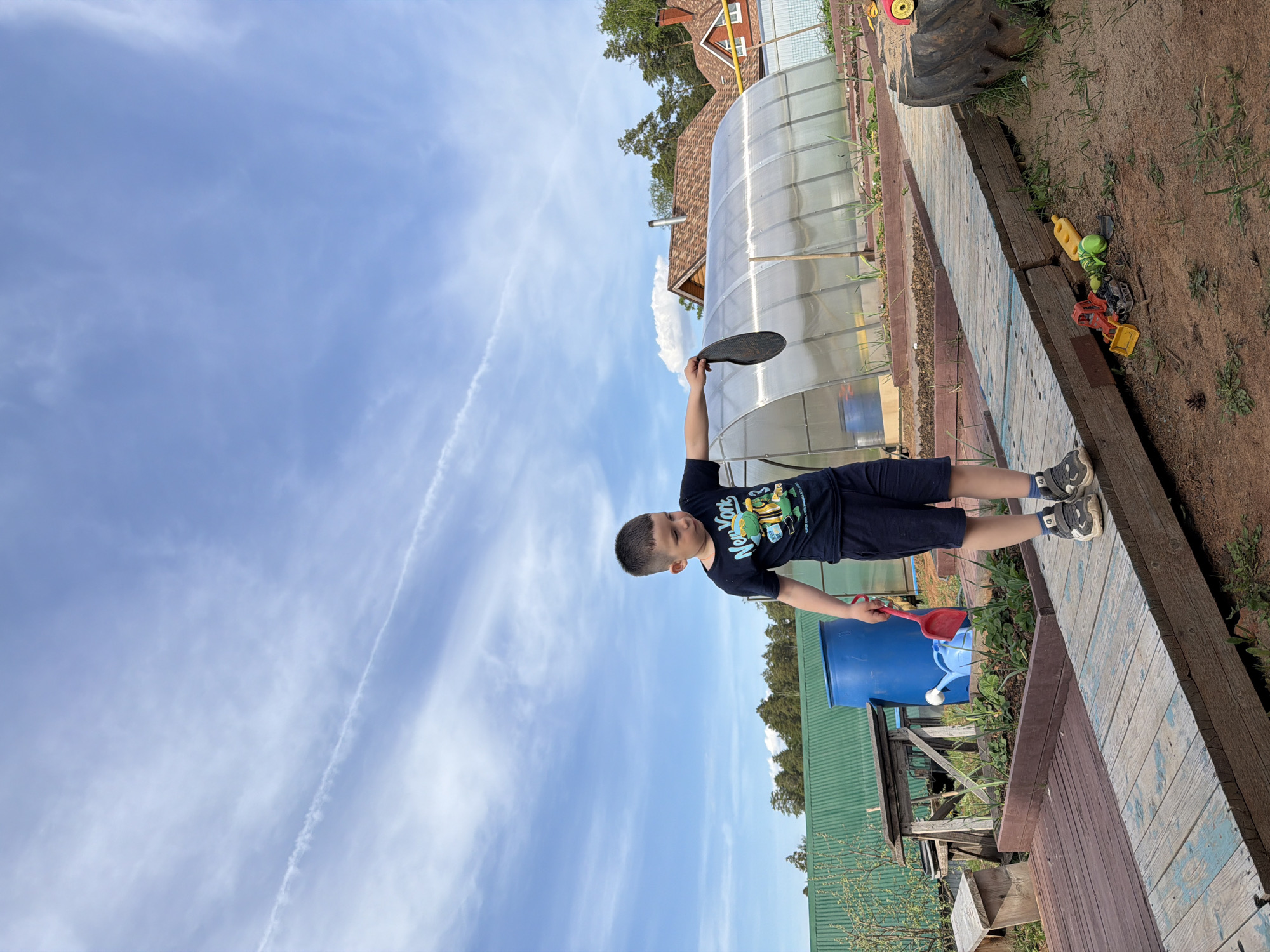 A young boy stands outdoors on a weathered wooden walkway adjacent to a garden area, holding a dark, flat object in his raised right hand, potentially a frying pan or something similar. He is wearing a dark blue t-shirt with a graphic on it and dark blue shorts. The setting is a backyard in what is likely a residential area. Behind the boy are various objects including a blue barrel, a red scoop, and what appears to be gardening equipment. Further back, a greenhouse and a glimpse of a house can be seen, along with green vegetation. The sky above is light blue with some wispy clouds and contrails. The perspective of the photograph has been rotated, making the sky appear to be to the left of the boy and the garden to the right. Considering the description of the location as Chaikovsky, Russia, the environmental cues such as the style of the buildings and vegetation align with this geographic region.