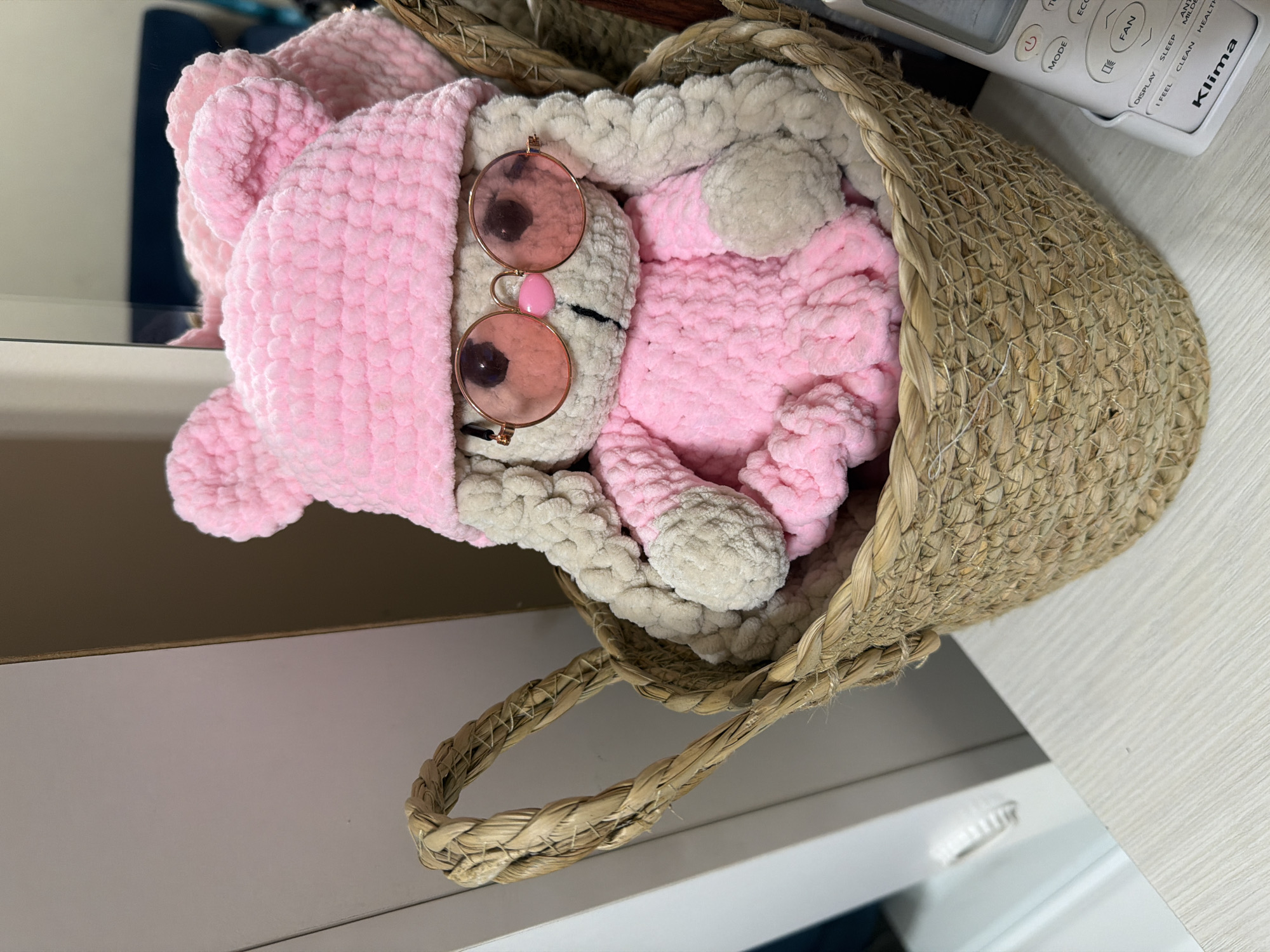A close-up, eye-level shot captures a handmade crochet bunny toy, predominantly pink and light beige, resting inside a woven straw basket. The bunny wears a pink crocheted hat with ear details and pink-tinted round sunglasses, obscuring its eyes. A small pink nose is visible below the sunglasses. The bunny is dressed in a pink crocheted outfit. The basket has two braided handles and is filled with the bunny. The background is softly blurred, showing a light-colored surface, possibly a table or shelf, and a hint of a wall or furniture. To the right of the basket, a white remote control with buttons and text, including the brand "Klima," is positioned. The overall lighting is bright, highlighting the textures of the crochet and basket. The image does not contain any elements that would suggest a location in the city of Shymkent, Kazakhstan.