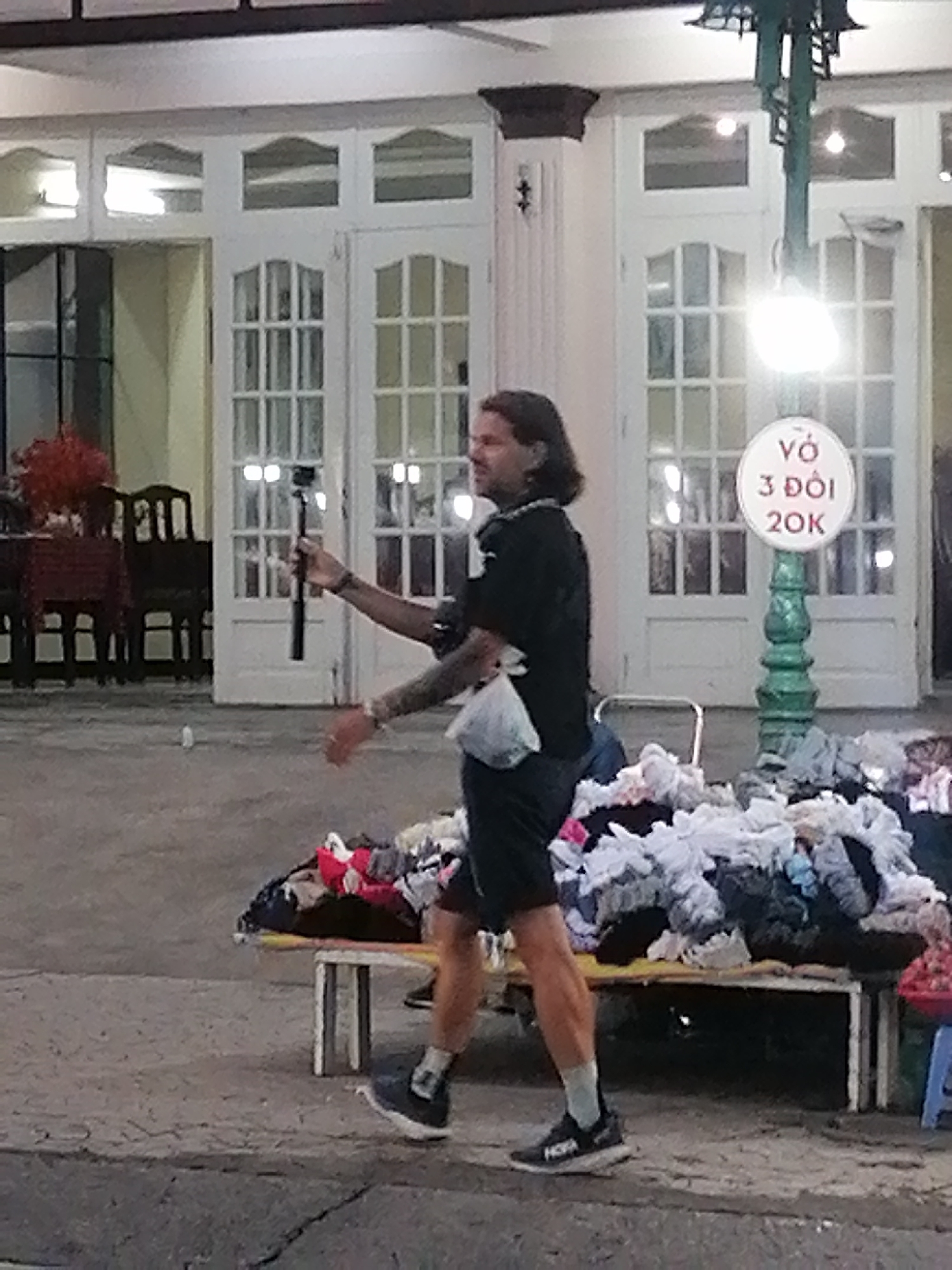 The image depicts a man in shorts and a black t-shirt holding a selfie stick and walking past a sidewalk vendor's wares in Da Lat, Vietnam. The vendor has a table piled high with clothes for sale. A sign on a green lamppost advertises "VO 3 DOI 20K," possibly indicating a price of 20,000 Vietnamese Dong for three items of clothing. Behind the vendor and the man are white, ornate double doors leading into a building. There are tables and chairs visible through the doors, and a red flower arrangement. The setting appears to be at dusk or in the evening, under artificial light.