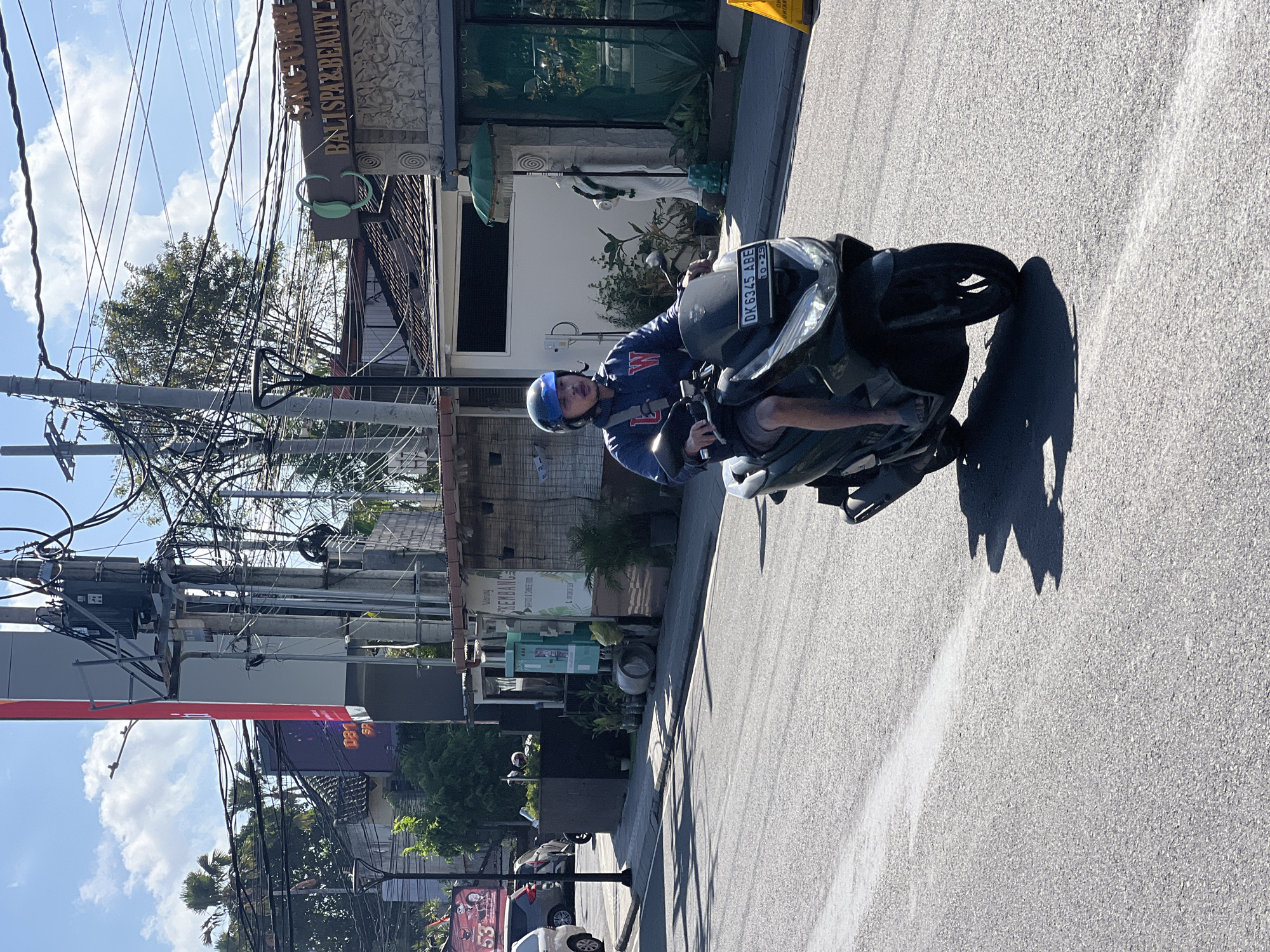 A man wearing a blue helmet and a blue and red jacket rides a black scooter down a paved road. He is wearing grey shorts and sandals. The scooter has a license plate that reads "DK 6345 ABE". The man is looking straight ahead. The scooter is casting a shadow on the road. In the background, there are buildings, power lines, and trees. One building has a sign that says "BALI SPA & BEAUTY". Another sign is red with white text and features the number "53". The sky is blue with some clouds. The scene appears to be in the city of Denpasar, Indonesia.