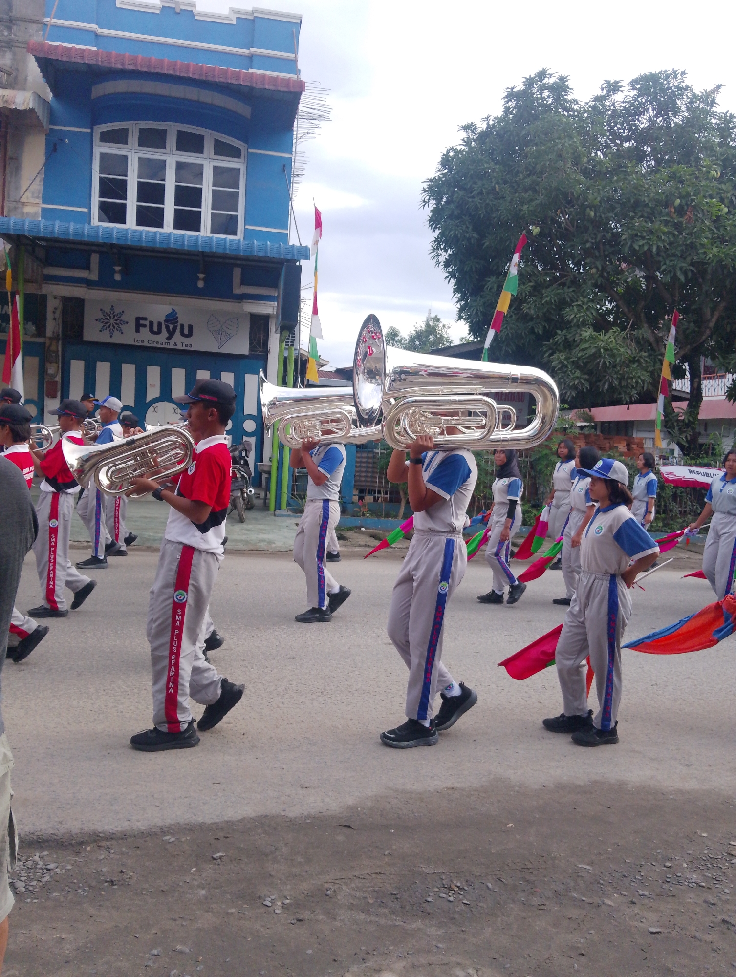 A marching band procession is taking place on a paved street in Serbelawan, Indonesia. The band members, predominantly young people, are dressed in matching gray uniforms with blue and red accents. Some are carrying large, silver brass instruments, specifically tubas, which are held horizontally over their shoulders. Others are holding colorful flags, red and blue, that are trailing behind them. In the background, a bright blue building with white trim and a red awning is visible, featuring a large multi-pane window and signage that reads "Fuyu Ice Cream & Tea". Trees with lush green foliage are also present, some adorned with red, white, and green flags. The sky is overcast. The ground is asphalt, with some gravel and debris along the side. A motorcycle is parked near the blue building.