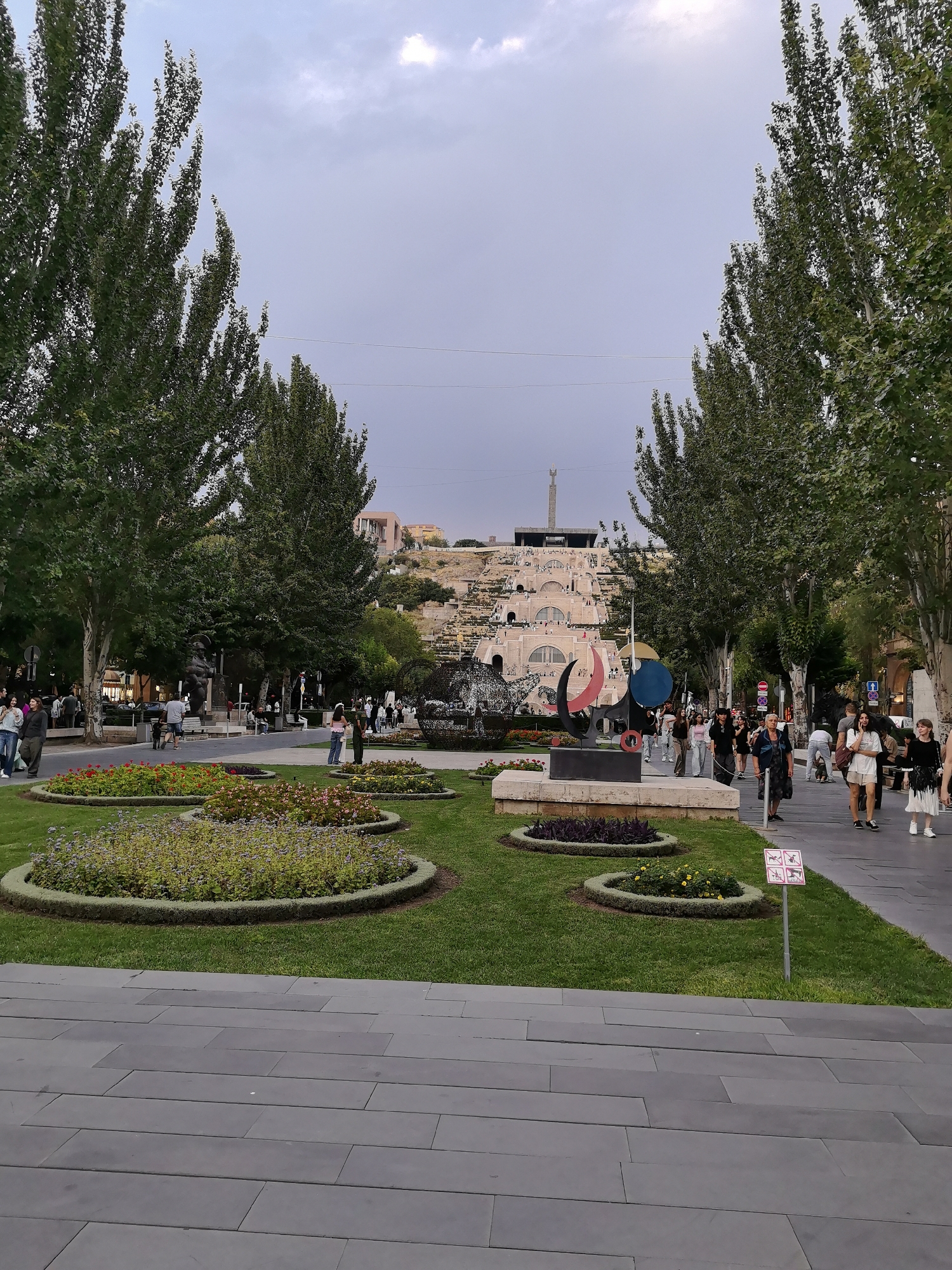 This image captures a view of the Cascade, a monumental staircase in Yerevan, Armenia, from the surrounding park area. In the foreground, manicured green lawns are interspersed with circular flower beds featuring various colorful blooms. A paved walkway runs along the right side of the image, with several pedestrians walking in both directions. To the left, tall trees with lush green foliage line the path.
In the midground, a striking abstract sculpture with curved shapes and bold colors stands prominently. Beyond the sculpture, the lower section of the Cascade, a massive limestone staircase, begins to ascend a hillside, featuring archways and a water feature. The upper portion of the Cascade culminates in a taller structure with a monument or obelisk at its peak. The background reveals more of the city's architecture and the sky, which is partly cloudy and overcast. People are visible walking on the Cascade stairs, suggesting it is a popular public space. The overall scene depicts a vibrant urban environment with artistic elements and recreational spaces.