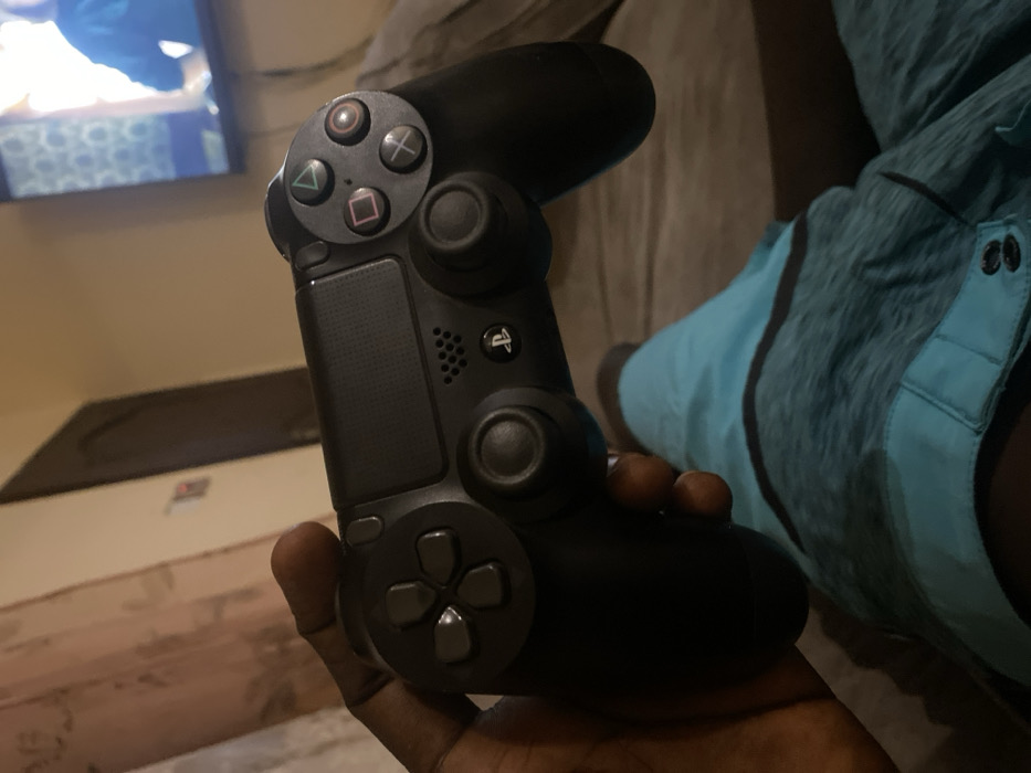 The image captures a close-up, angled view of a person's hand holding a black PlayStation DualShock 4 controller, set indoors in Lakka, Sierra Leone. The dark-skinned hand firmly grips the controller, with fingers visible wrapped around the bottom.
The controller is the primary focus, showcasing its distinct features. On the left side, the iconic face buttons—triangle, circle, cross (X), and square—are clearly visible, along with the directional pad below them. The two analog sticks are also prominent, with the left one partially obscured by the hand. The central touch pad, featuring the PlayStation logo (PS button) and a small speaker grille, is also distinct.
In the blurred background, a television screen displays bright, indistinct content, suggesting a video game or other media is being played or watched. To the right, part of a leg wearing blue and teal athletic shorts or sleepwear is visible, indicating the person is likely relaxing on a couch or bed. The overall lighting is dim, suggesting an evening or nighttime setting. The scene conveys a relaxed atmosphere of leisure and entertainment.