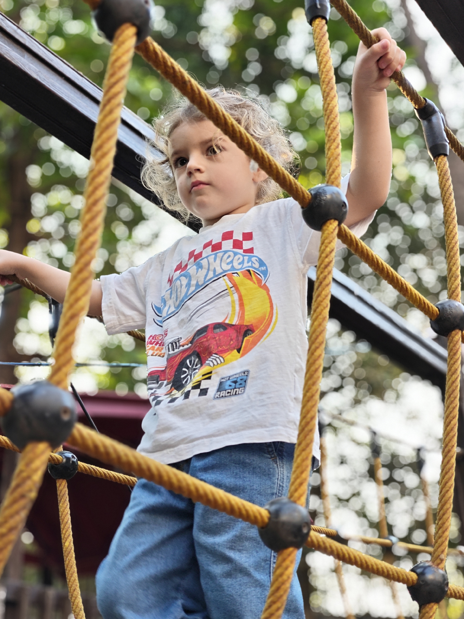 A young child, approximately 3-6 years old, with light, curly hair, is depicted standing on a yellow-orange rope climbing net within an outdoor playground in Mumbai, India. The child wears a white short-sleeved t-shirt featuring a "Hot Wheels" graphic with a red race car and checkered flag pattern, along with blue denim jeans. The child's right hand firmly grasps a vertical section of the rope net above their head, while the left arm extends horizontally to the left, also in contact with the structure. The child's head is slightly tilted to the left, with eyes directed upwards and to the left. Behind the child, a dark structural beam of the climbing equipment is visible. The background is blurred, showing green foliage indicative of trees and vegetation, under bright daylight conditions.