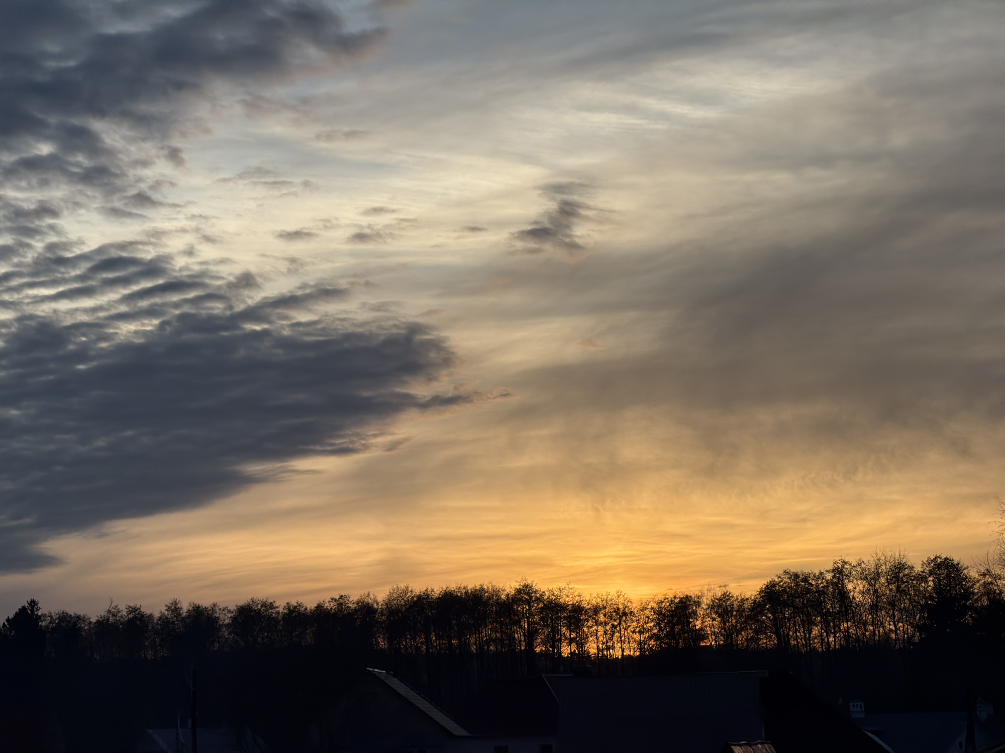 The scene, located in Chaikovsky, Russia, depicts either a sunrise or sunset. The upper two-thirds of the image display a sky with a gradient of atmospheric conditions, transitioning from dense, dark grey cumuliform clouds in the upper left to a expanse of lighter grey, white, and vibrant orange-yellow hues across the center and right, where the sun's light is most prominent, though the solar disk itself is not visible. A distinct, dark silhouette of a treeline extends horizontally across the lower middle section of the frame, with the bare or sparsely leaved branches of individual trees faintly discernible against the illuminated sky. Below the treeline, in the foreground, are the dark, indistinct silhouettes of multiple building rooftops, including a prominent triangular gable roof on the lower left. No individuals or discernible activities are present within the captured environment.