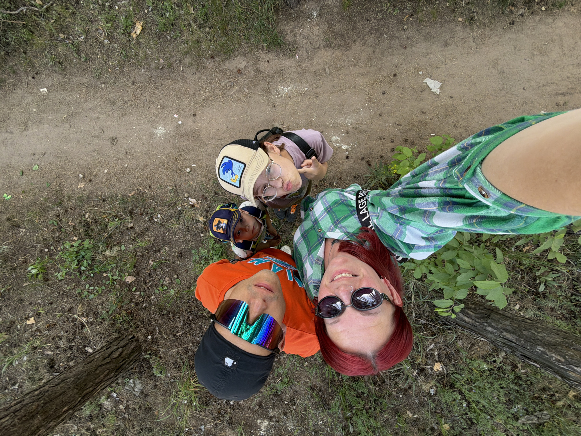 Three adults and one child are visible in an upward-angle selfie taken outdoors. The setting is a wooded area in the city of Chaikovsky, Russia. The ground is covered with sparse grass, dirt, and some fallen leaves. A dirt path is visible at the top of the frame. A log lies to the right.
The person holding the camera has red hair, is wearing sunglasses, and a green plaid shirt. An adult man is wearing an orange shirt, black baseball cap, and iridescent sunglasses. An older child wears a hat with a Road Runner cartoon character, glasses, and a backpack. A younger child wears a hat with an unknown design. All are looking up at the camera.