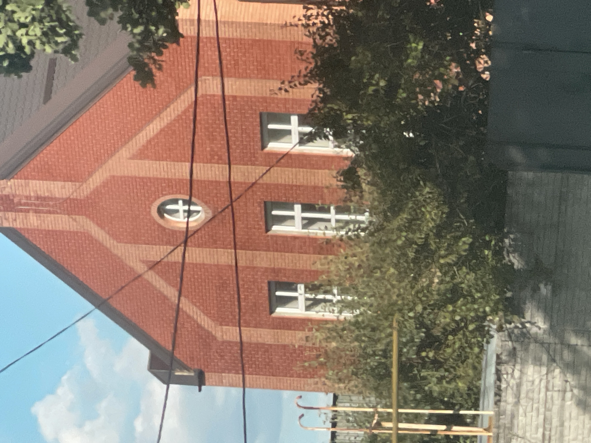 The image shows the facade of a red brick building, possibly a church or a similar structure, in Poltava, Ukraine. The building has a peaked roof and several windows. One circular window with a cross-like element is visible near the apex of the roof. Below this, three rectangular windows with white frames and crossbar dividers are situated. Dense green foliage obscures the lower portion of the building. In the foreground, a low brick wall and a dark fence are visible. Overhead, power lines crisscross the sky, which is partly cloudy. The scene appears to be a street view, with no people or vehicles present.