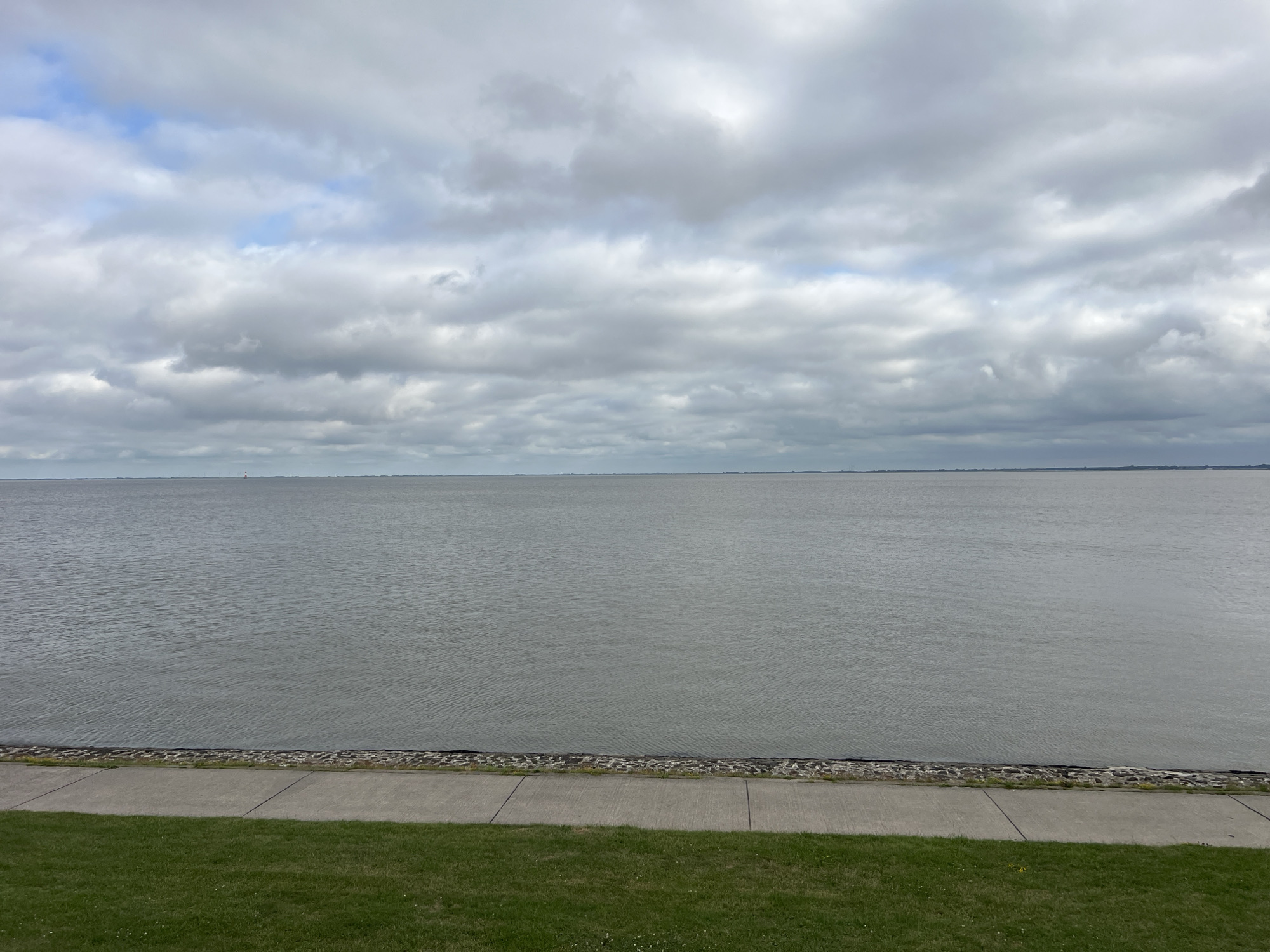 The image depicts a wide, calm body of water under a cloudy sky. In the foreground, there is a strip of green grass, followed by a paved walkway. Beyond the walkway is a sea wall made of grey stones. The water extends to the horizon, where a faint line of land and a small structure, possibly a lighthouse, can be seen. The overall impression is one of a serene, coastal landscape. This view is consistent with the Jade Bight in Wilhelmshaven, Germany, a large bay known for its calm waters and coastal paths. There are no people or animals visible in the image.