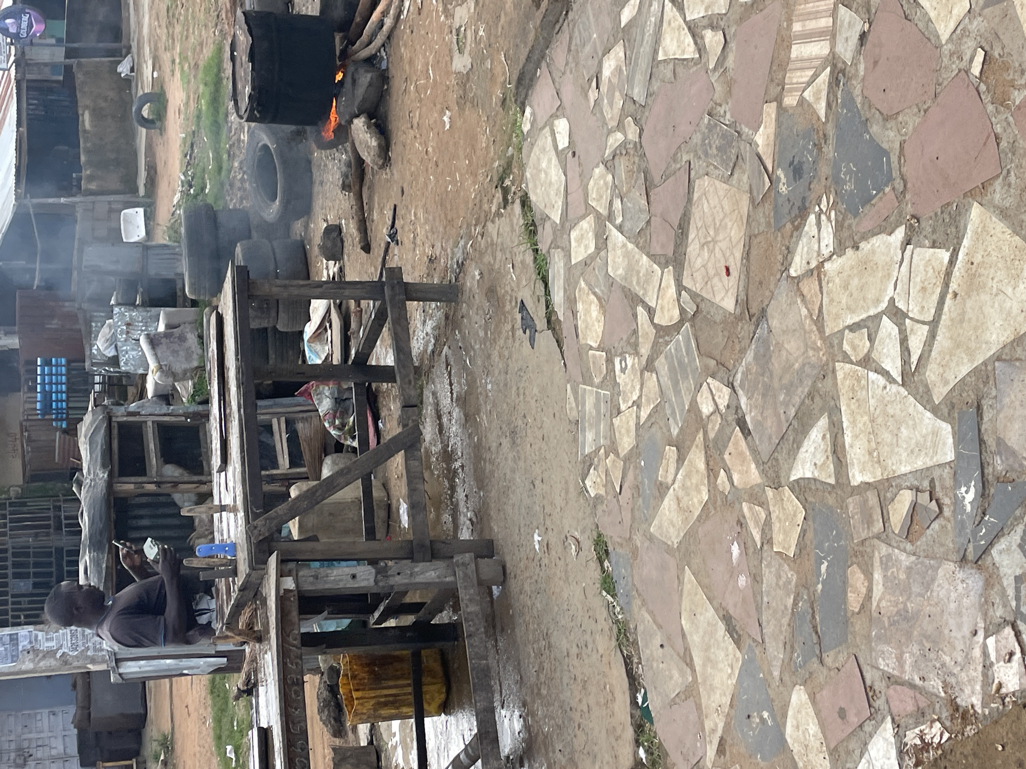 The image depicts an outdoor scene in Rafin Par, Nigeria. In the foreground, a patterned stone patio is visible. To the left, a man in a dark t-shirt is standing and looking at a mobile phone, holding some items in his other hand. He is positioned behind a wooden table setup. Further into the scene, there are wooden structures, tires stacked and scattered, and a large barrel on a fire that is producing smoke. The ground is a mixture of dirt and debris. The overall environment suggests a marketplace or an informal commercial area.
