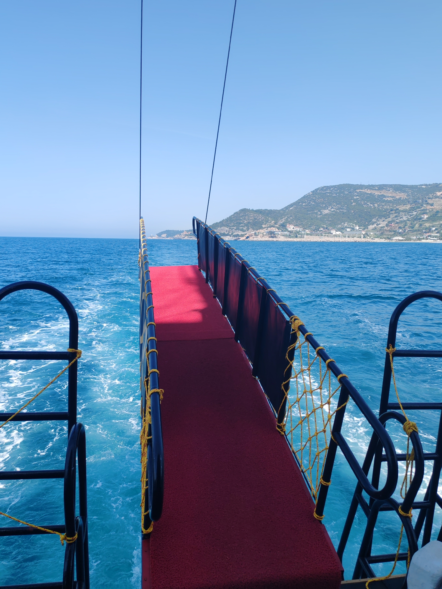 The image depicts a view from the rear of a boat moving through the sea, likely near Alanya, Turkey, as suggested by the visible coastline and landscape in the background. The boat has a red-carpeted platform extending towards the water, enclosed by black metal railings. Yellow rope details are visible along the railings. The sea is blue and foamy, stirred up by the boat's movement. In the background, a green hill or mountain is visible, dotted with buildings. The sky is a clear, light blue. The presence of the red carpet suggests a recreational vessel, possibly used for tourism.