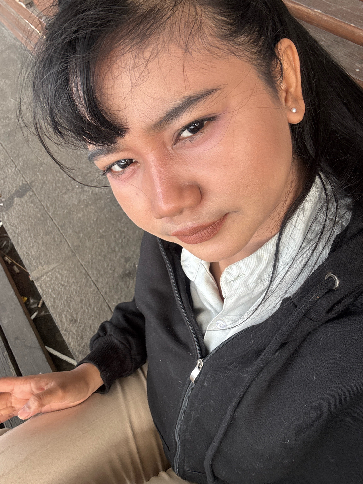 A medium-shot, high-angle, selfie-style photograph shows a young person of Indonesian ethnicity with dark hair and makeup posing on a bench outdoors. The person is wearing a black hooded jacket over a light blue collared shirt and light-colored pants. Their left hand rests on their lap. The background consists of a grey, textured surface, likely a paved area with faint lines, and the dark slats of a bench. A small, white pearl earring is visible in the person's right ear. The lighting suggests an overcast day. The specific location in North Jakarta, Indonesia is not discernible from the visual information provided.