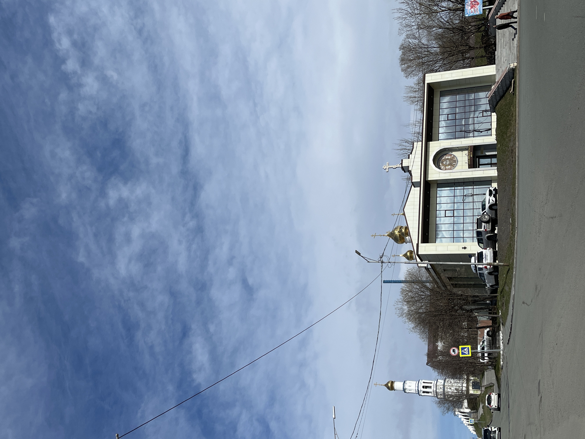 The image depicts a street scene in Petropavlovsk-Kamchatsky, Russia. A modern church with a white facade and a combination of traditional domes and modern architectural elements is prominent on the right side of the frame. The building has large windows that reflect the sky, and a cross is visible on its roof. To the right of the church is a parking lot with several vehicles. Adjacent to the church is a sidewalk, beyond which two individuals are visible. In the distance, a traditional white bell tower with gold domes rises above the trees. Streetlights and power lines are also visible in the composition. A pedestrian crossing sign is seen near the tower. The sky occupies a significant portion of the image and is filled with blue sky and white clouds.