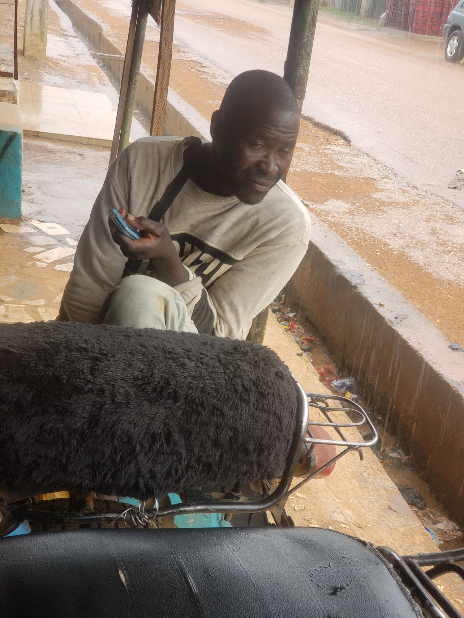 A Black man is sitting on a motorcycle in Sabon Tasha, Nigeria, holding a cell phone and looking to the right. He is wearing a light-colored long-sleeved shirt with a black design across the chest and light-colored pants. The man is seated under a shelter with a wooden post and metal roof. The ground is wet, indicating it has been raining. In the background, there is a gutter filled with trash and debris, and a street with a vehicle visible in the distance. The motorcycle has a dark-colored furry seat cover and a metal rack behind it.