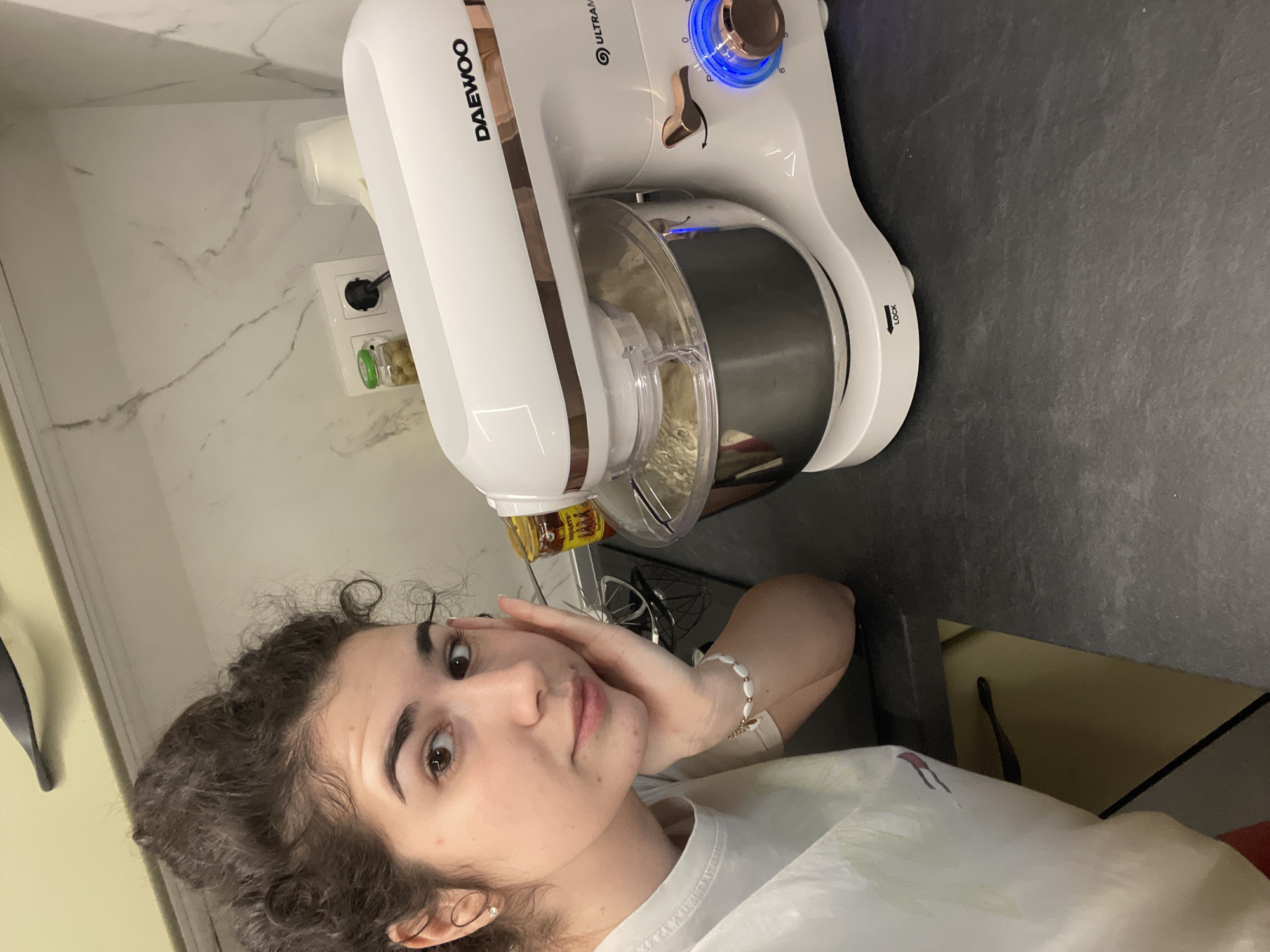 A young woman with curly hair is posing for a selfie in what appears to be a kitchen. To her upper right is a white and rose-gold Daewoo stand mixer on a dark gray countertop. The mixer is plugged into an electrical outlet on a white marble wall. Inside the mixing bowl, there is a light-colored substance. A bottle, possibly of vanilla extract, is visible next to the mixer. The location in Sofia, Bulgaria cannot be determined from the image.