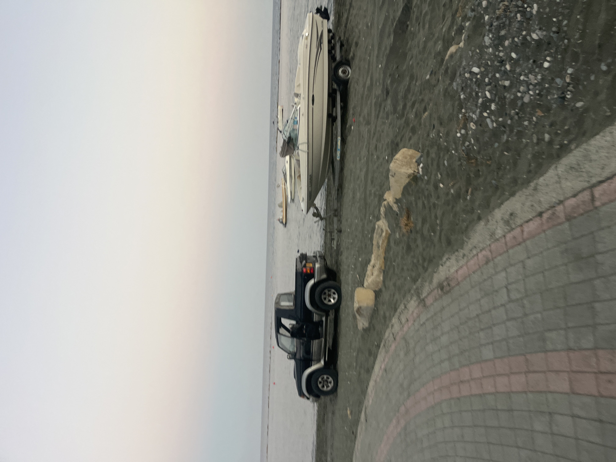A black pickup truck is parked on a dark sandy beach near the water's edge. The driver's side door is open. Behind the truck, a light-colored speed boat on a trailer is also positioned on the beach, facing the water. In the distance, the calm sea meets a pale, hazy sky. Further out on the water, other boats are visible. The foreground shows a paved walkway with a patterned surface of grey and pinkish-red tiles. Rocks and some sparse vegetation are scattered on the sand. The scene appears to be in Agios Tychona Community, Cyprus, given the context of the prompt.