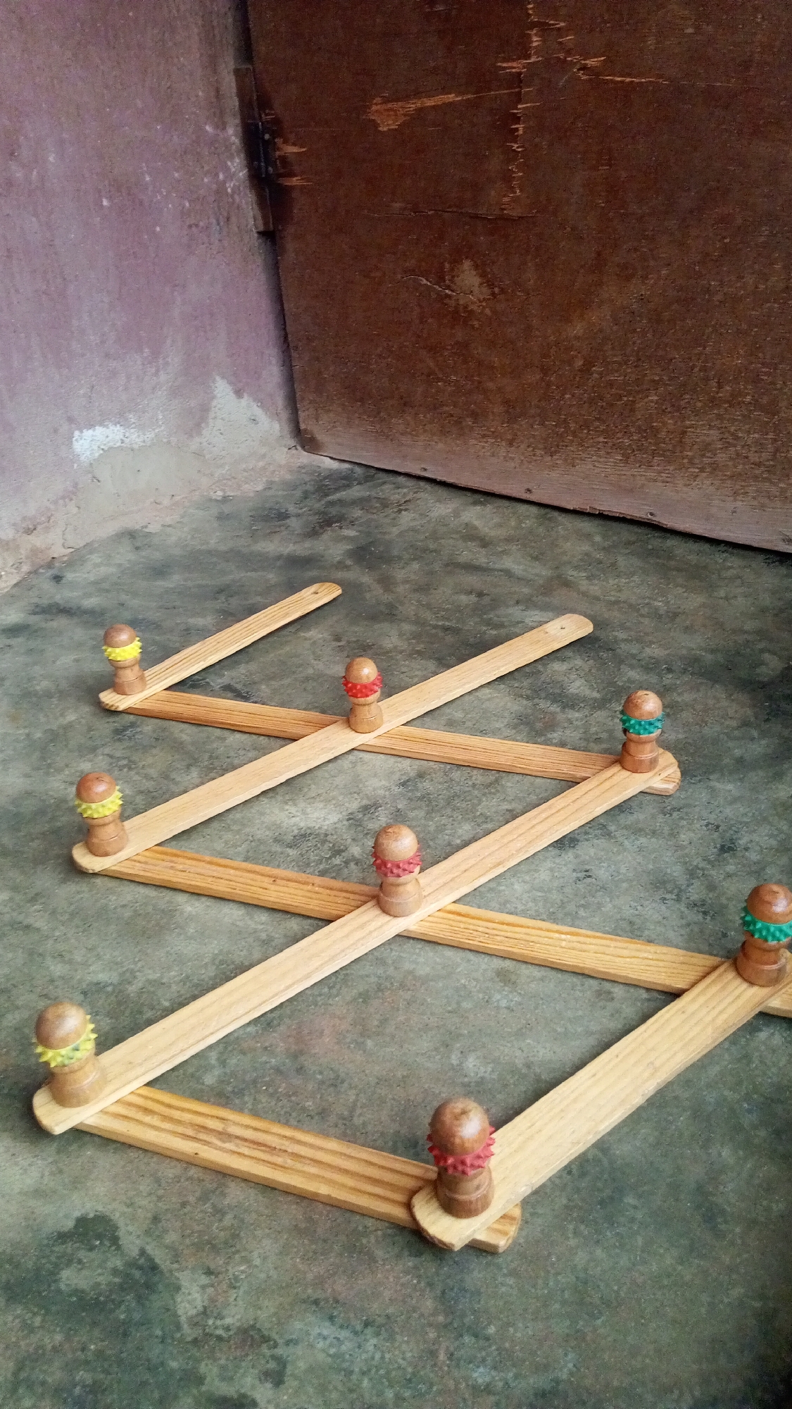 A wooden coat rack lies on a mottled gray concrete floor in front of a brown wooden door and a painted wall in a possible interior space. The coat rack has a lattice structure made of light-colored wooden slats with wooden knobs topped with colorful accents: yellow, red, green. The door shows visible wear and tear. The scene appears to be located in Onitsha, Nigeria.