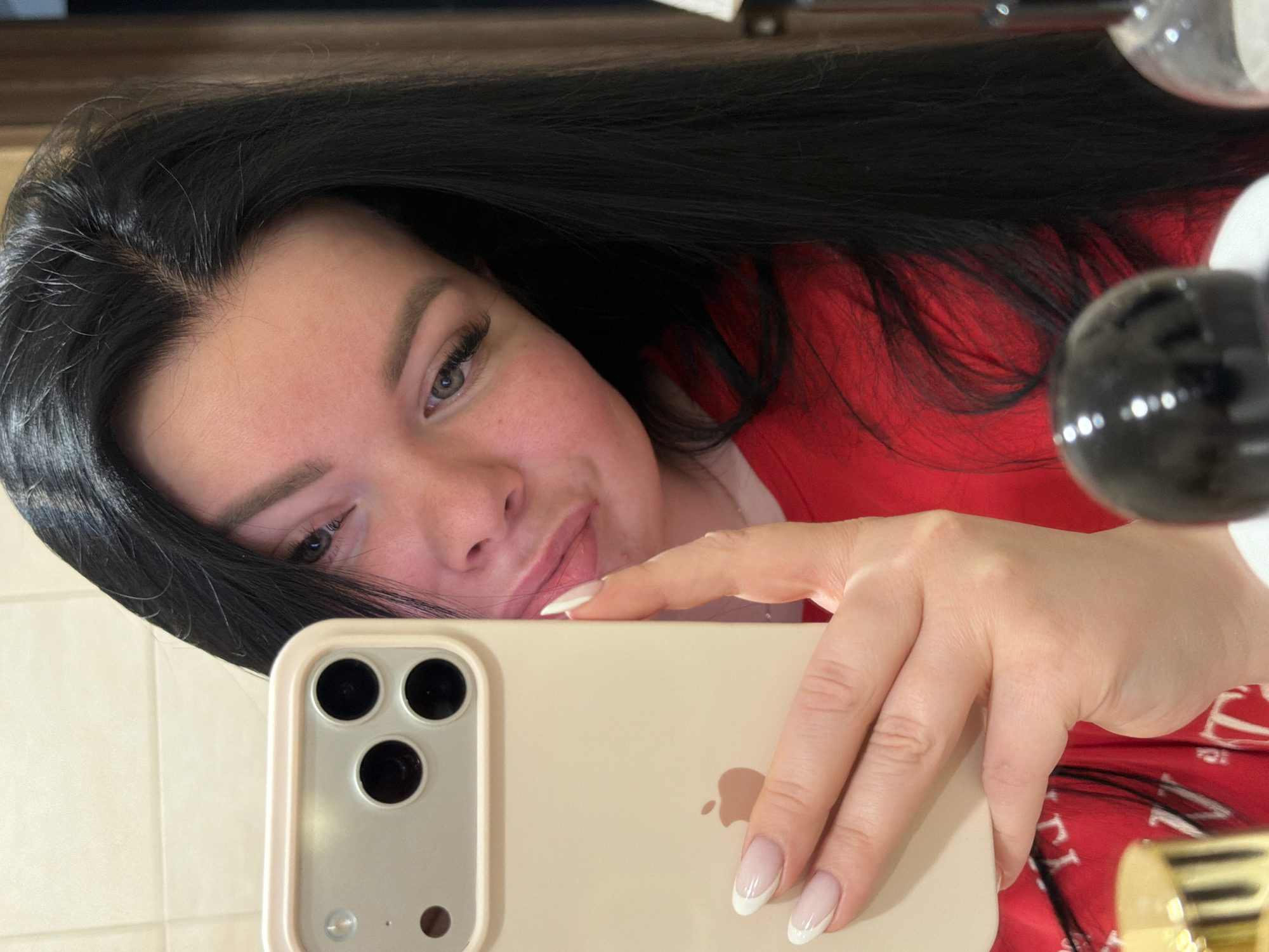 A person with long, dark hair takes a selfie in a mirror with an iPhone. The phone has a beige case and is held in their right hand, displaying French manicured nails. The person is wearing a red garment. The location is not identifiable as Chaikovsky, Russia based on the image content.