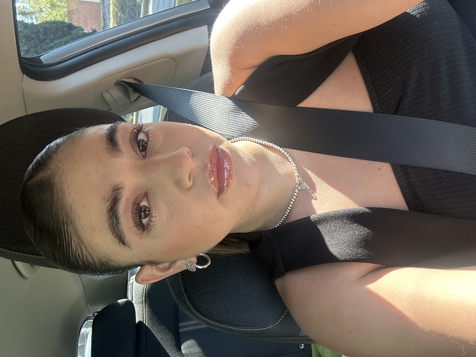 A young woman wearing a black hat, black top, seatbelt, and silver necklace is seated in a car. She is wearing earrings and has glossy lips. The car is likely in Puebla, Mexico, based on the general visual style of the vehicle and the building visible through the window. The woman is looking at the camera. Sunlight is visible in the scene.