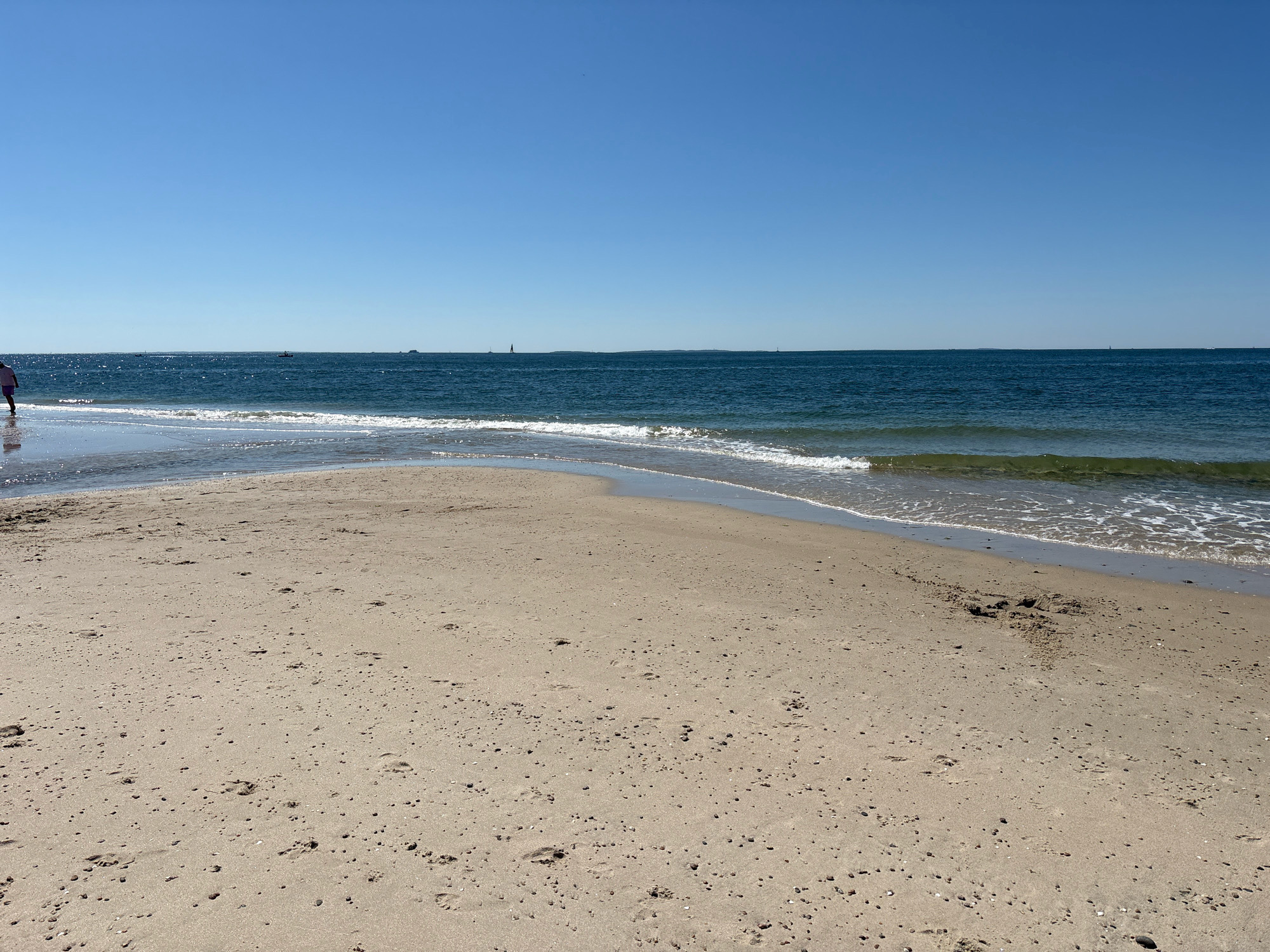This is an eye-level, wide shot of a sandy beach on a clear, sunny day. The sand is light brown and speckled with small shells and pebbles. There are numerous footprints pressed into the sand, indicating recent human activity. In the background, the deep blue ocean meets the horizon under a bright, cloudless blue sky. Gentle waves are lapping at the shore, creating a thin line of white foam. A solitary person, seen from behind and partially cropped, is walking along the water's edge on the left side of the frame. The person is wearing pink shorts and a light-colored top. In the distance, several small boats and a larger ship are visible on the ocean. The scene evokes a peaceful and serene coastal environment. The location is a beach in Waterford, United States of America.