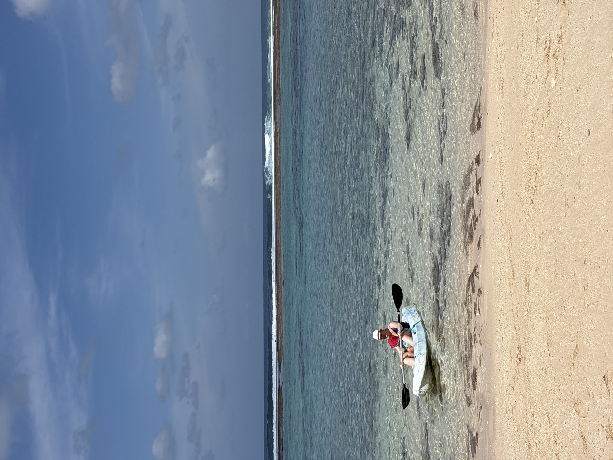 A person is seated in a kayak near the shore of Kuta Selatan, Indonesia. The kayak is light blue and white with a black paddle extended across it. The person is wearing a white hat, sunglasses, and a dark swimsuit. They are holding the paddle with both hands, poised to move through the water. The water is clear and shallow, revealing rocks and coral formations beneath the surface. In the background, a reef breaks the turquoise water, with waves crashing beyond it. The sky is a bright blue, dotted with scattered white clouds. The foreground shows a sandy beach with small pebbles and shells.