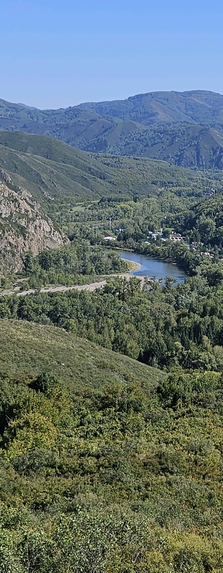 The image is a vertical, high-angle shot of a lush green valley with a winding river and a small settlement. In the foreground, the dense foliage of trees and shrubs covers a sloping hillside. The middle ground features a wider valley floor with a river snaking through it. On the far bank of the river, a cluster of buildings, likely houses, forms a small village, interspersed with trees. The landscape is framed by rolling, forested hills that recede into the background, with a clear blue sky overhead. The location is identified as the Tarkhanka rural district in Kazakhstan. The scene depicts a tranquil, rural environment characterized by natural beauty and a human settlement integrated within it. No people or specific actions are visible.