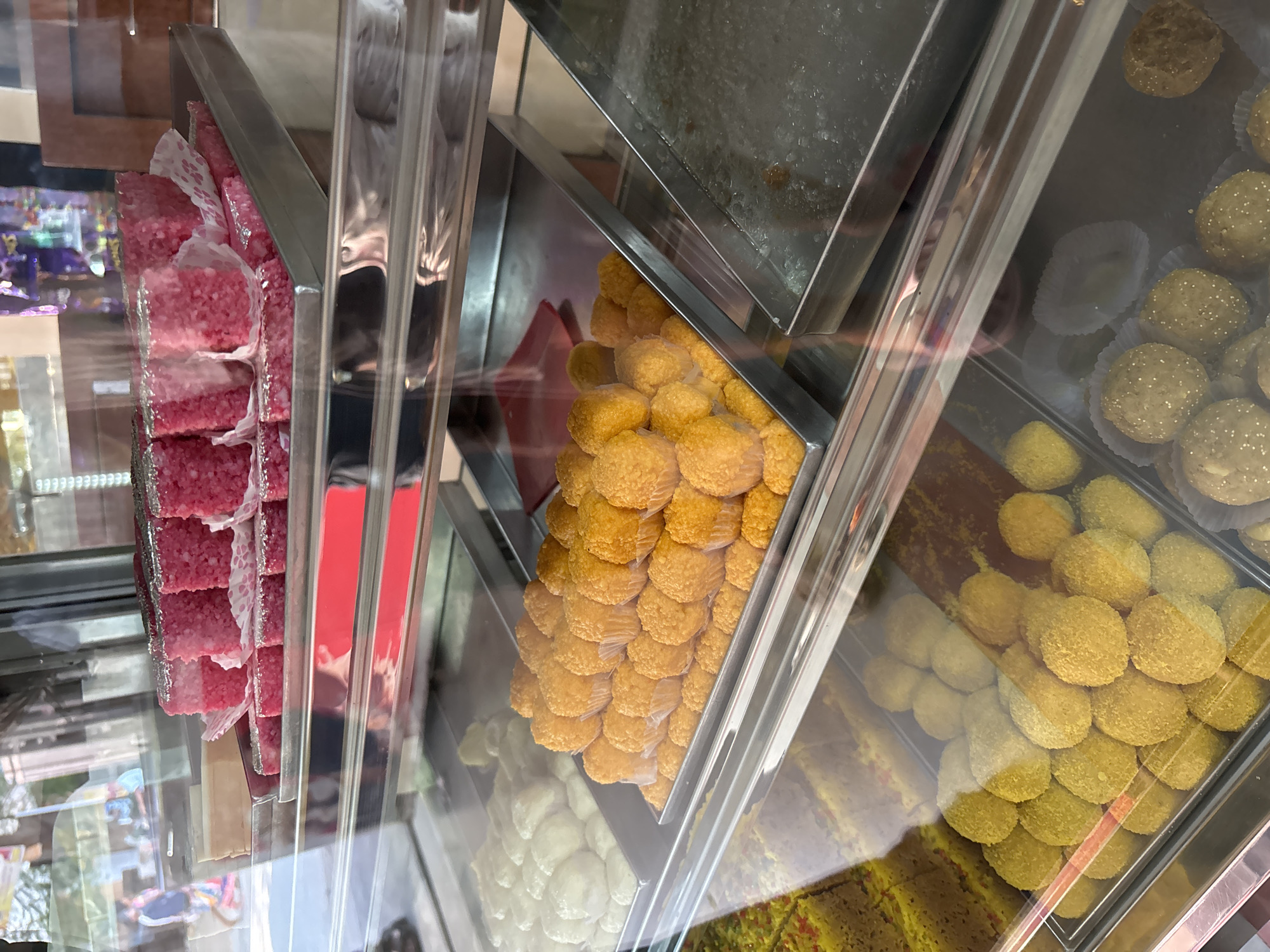 Inside a glass display case, arranged in vertical rows within metal trays, are several varieties of Indian sweets. The leftmost tray holds pink, cube-shaped sweets, separated by white paper. The tray next to it contains round, yellow sweets densely packed. Further to the right, there are trays filled with round, yellow sweets, interspersed with yellow, bar-shaped sweets. At the top right, round, light-brown sweets are visible inside white paper liners. The overall scene is a close-up view of a sweet shop's display, likely in Ambernath, India, based on the type of sweets shown.