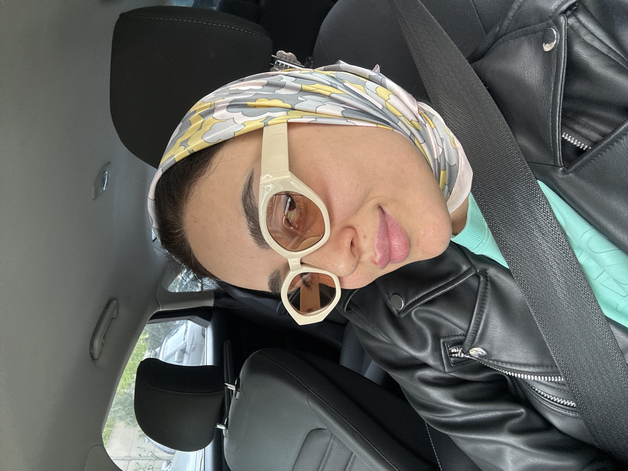 A person is inside a car, wearing a headscarf and sunglasses, and secured with a seatbelt. They are wearing a black leather jacket over a light blue top. The car interior features gray upholstery and a black headrest. The person's face is prominent in the frame. Outside, a vehicle and some greenery can be seen through the car window. The location cannot be determined from this image alone.