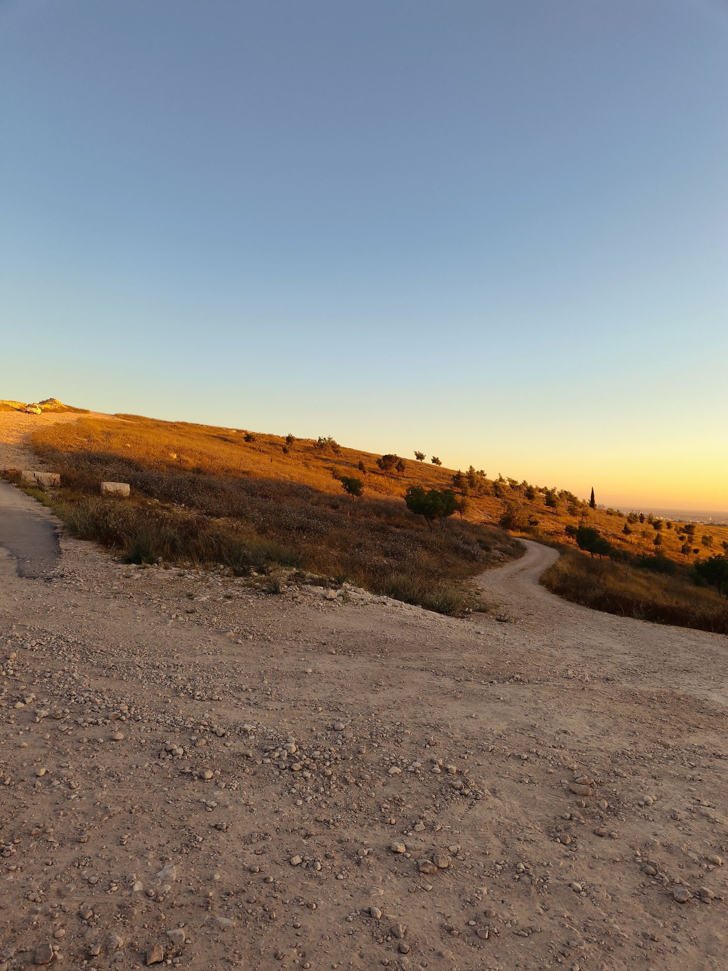 The image depicts a dirt path winding up a gently sloping hillside in Be'er-Sheva, Israel, during what appears to be sunset. The sky transitions from a clear, light blue at the top to a warm, golden orange near the horizon. The hillside is covered in dry, golden-brown vegetation, interspersed with sparse trees and shrubs. Several scattered, light-colored rocks are visible along the path and on the hillside. In the distance, a hazy view of a city can be seen. The path itself is composed of dirt and small stones, with some tire tracks visible in the foreground. The overall impression is one of a rural or semi-rural landscape bathed in the warm light of the setting sun.
