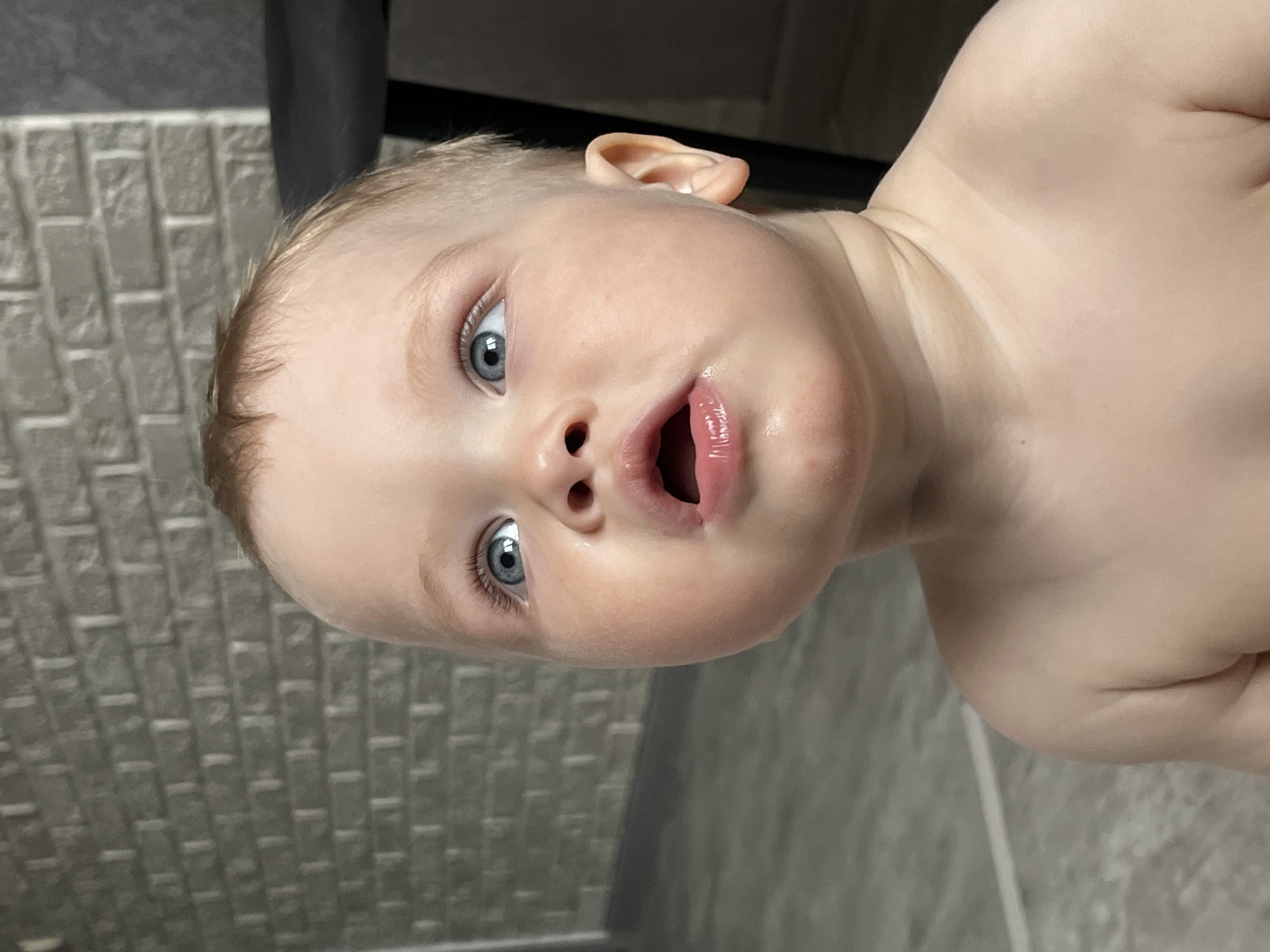 A close-up, slightly tilted image captures a young child with fair skin, blond hair, and striking blue eyes. The child's mouth is open, giving a surprised or inquisitive expression. The background features a textured wall resembling brickwork on the left and a plain, light-colored wall on the right, with a dark shape partially visible at the top. There is no definitive evidence to confirm that the picture was taken in Chaikovsky, Russia, or anywhere else for that matter.
