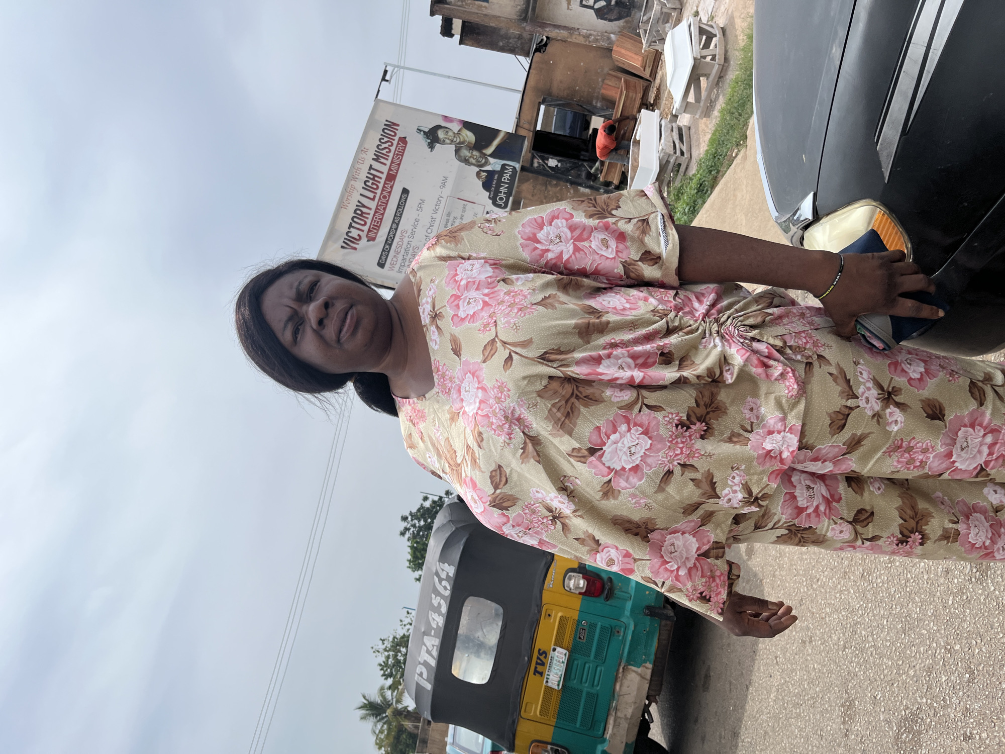 A medium shot captures a Black woman standing outdoors, facing the camera. She wears a beige and pink floral pajama set. Her dark hair is pulled back, and her expression is neutral. To her left, a green and yellow auto-rickshaw with a black canopy is visible. The auto-rickshaw's license plate reads "PTA-4564" and "BLU-560A." In the background, a large banner reads "VICTORY LIGHT MISSION INTERNATIONAL MINISTRY," with text below detailing worship services. A man in a red shirt is seen working near a collection of caskets of various sizes and colors. The scene appears to be on a street in Jos, Nigeria, with a cloudy sky overhead and some greenery visible. A black car is partially visible in the foreground on the right side of the image.