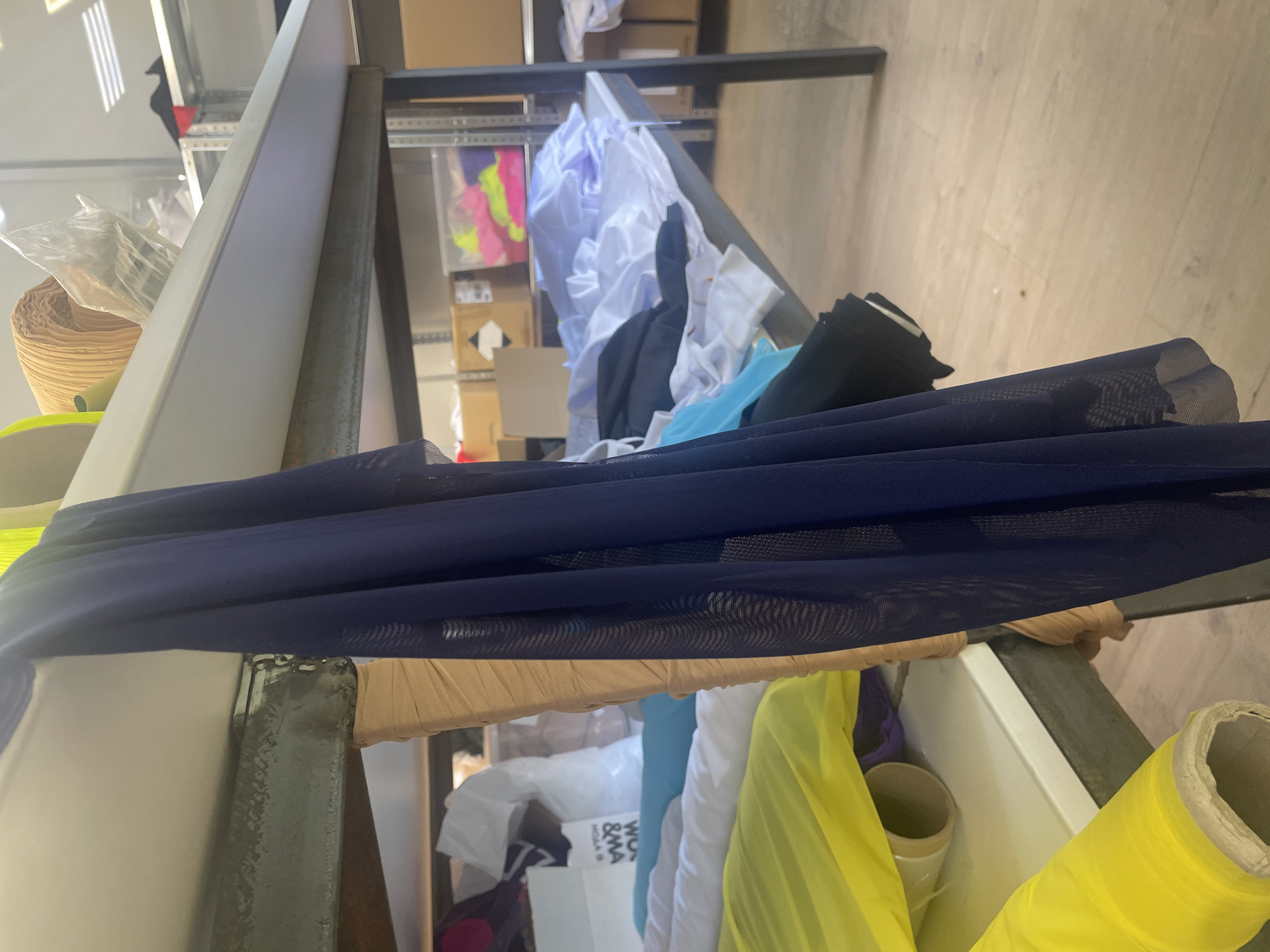 The image depicts a storage area, likely within a textile workshop or warehouse, filled with rolls of fabric. The scene is brightly lit by overhead fluorescent lights, reflecting off the smooth, light-colored surfaces of the shelves and the wooden floor. In the foreground, a dark blue mesh fabric hangs prominently, partially obscuring the view of other materials. These include rolls of yellow and tan fabric on lower shelves, and stacks of white and light blue fabric on an upper shelf. The shelves themselves are constructed of metal, with a worn, industrial appearance. In the background, more shelves are visible, holding cardboard boxes and plastic containers filled with brightly colored fabrics, including neon pink and yellow. The overall impression is one of a busy, functional space dedicated to the storage and organization of textiles. There is no indication of the location being in Saint Petersburg, Russia.