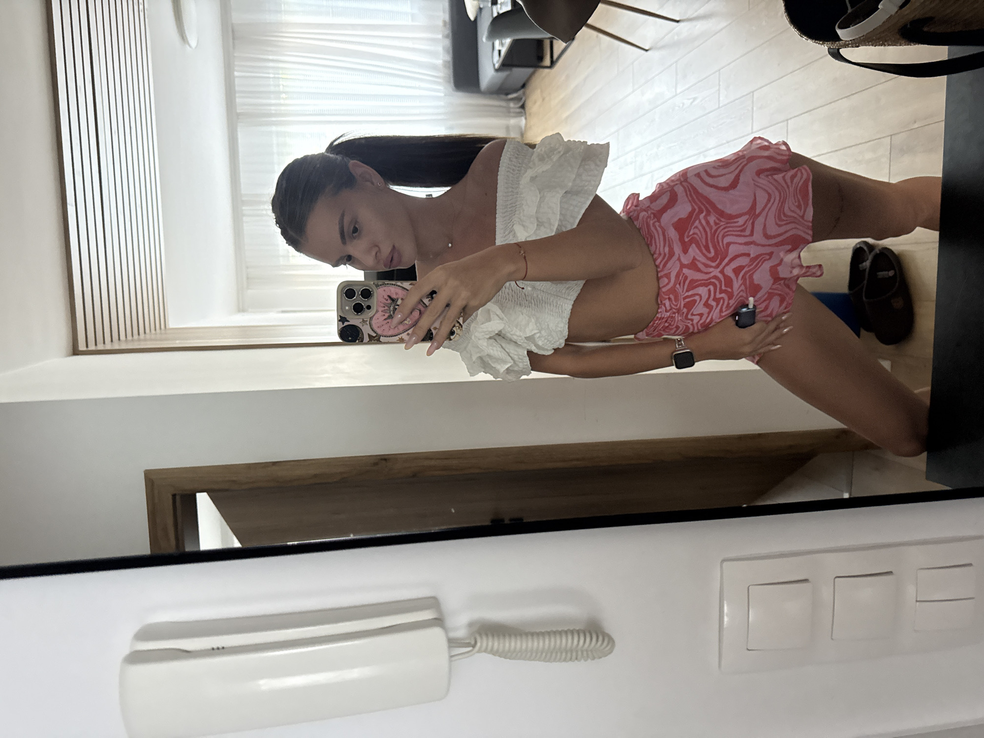 A young woman is taking a mirror selfie in a modern room. She is wearing a white off-the-shoulder crop top with ruffled sleeves and pink and red swirled pattern shorts. Her hair is pulled back in a high ponytail. The background shows a doorway, a wall with a white intercom system and light switches, and a living area with furniture and sheer curtains. The flooring is light wood.