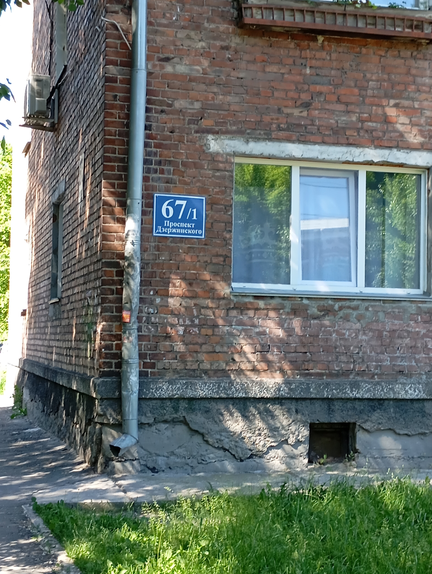 The image shows the corner of a brick building in Novosibirsk, Russia. A blue street sign with white text is affixed to a drainpipe on the corner. The sign reads "67/1 Проспект Дзержинского" which translates to "67/1 Dzerzhinsky Avenue". The building has a large white-framed window on the right side. The lower part of the building is made of concrete and stone, with some visible damage and wear. There is a small, dark opening in the concrete foundation. Lush green grass grows in front of the building and on the left side. The scene is lit by natural sunlight, casting shadows on the brickwork.