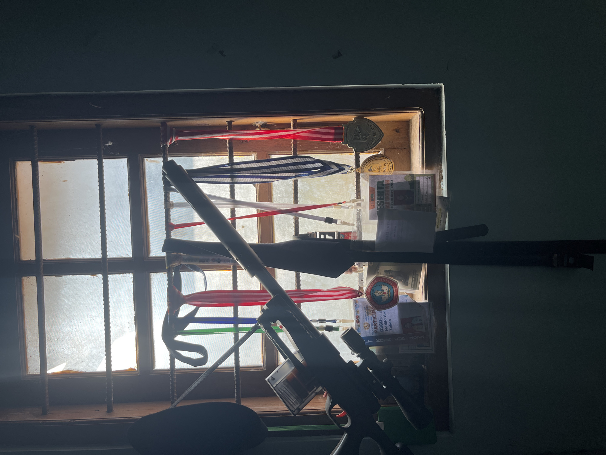 The image depicts a dimly lit interior scene focused on a window with several items hanging on bars in front of it. The window has multiple panes, some of which appear dusty or frosted, and sunlight is filtering through, creating bright patches. Hanging from the bars are several colorful ribbons of varying widths, some with medals attached. There are also some papers or certificates pinned to the bars. In the foreground, a dark object, which appears to be a disassembled rifle or air rifle, is positioned diagonally across the frame, obscuring part of the window and the items hanging on the bars. The room appears to be a private space, possibly a bedroom or study, given the personal items displayed. There are no people visible in the image. The overall impression is one of a personal display area, perhaps a trophy or achievement wall, within a private residence. There is no discernible information in the image that would indicate a specific location in Glagah Kulon, Indonesia.