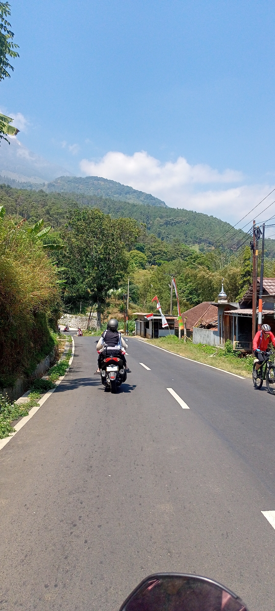 In Cembor, Indonesia, a road curves gently uphill amidst lush greenery. The sky is a vibrant blue, dotted with white clouds. On the right side of the road, a cyclist in a red jacket and helmet pedals forward, their bicycle reflecting the sunlight. Further ahead on the left, a motorcycle with two riders, both wearing helmets, travels up the incline. In the background, a verdant mountainside rises, covered in trees and mist. The road is paved and marked with white lines. Along the sides of the road, there are small buildings and vegetation, including bamboo and banana trees. Several Indonesian flags are visible, indicating a festive or patriotic atmosphere. The scene captures a moment of everyday life and transportation in a scenic, natural setting.