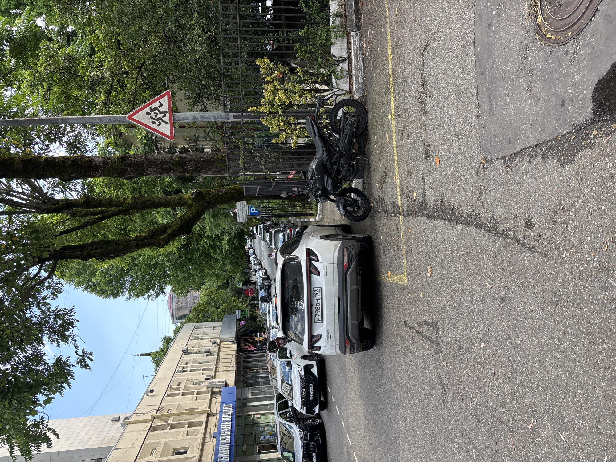 This media file shows a street scene in Sochi, Russia. The perspective is from a low angle, looking up and across the street.
In the foreground, a black motorcycle is parked on the side of the road, leaning on its kickstand. The road itself is asphalt with a yellow line marking the lane. To the left of the motorcycle, there is a white sign with a red border, depicting stylized figures of people running, indicating a pedestrian crossing or a warning about pedestrian activity. Further down the street, a white SUV is parked, followed by a line of other cars. The buildings lining the street are multi-story structures, with some featuring balconies and air conditioning units. One building has a blue sign with Russian text.
Behind the motorcycle and extending down the street is a dark metal fence, behind which are lush green trees and some lighter green foliage. The trees cast shadows on the road and sidewalk. Above, the sky is a bright blue, suggesting daytime. The overall impression is of a typical urban street, with parked vehicles and roadside infrastructure, set against a backdrop of dense greenery. The presence of the pedestrian warning sign and the parked cars suggest a functional urban environment. The bright sky indicates it is likely daytime, possibly late morning or early afternoon, and the greenery suggests a warm climate.
Visible text includes "P798BM 193" on the license plate of the white SUV. On the blue sign on the building, part of the text is visible and appears to be in Cyrillic script, with the word "БАНК" (Bank) clearly discernible.