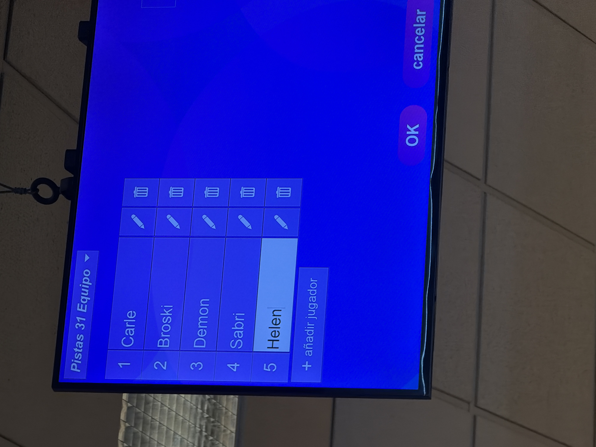 A digital display screen is angled upwards, showcasing a player management interface on a bright blue background. The interface header reads "Pistas 31 Equipo" (Lanes 31 Team), suggesting a bowling alley or similar recreational facility in the city of Puebla, Mexico. Five players are listed: 1. Carle, 2. Broski, 3. Demon, 4. Sabri, and 5. Helen. Each player name is accompanied by a white pencil icon (edit) and a white trash bin icon (delete). The entry for "Helen" is highlighted in white. Below the player list, a button labeled "+ añadir jugador" (add player) is present. At the bottom right, two buttons are visible: "OK" on a pink background and "cancelar" (cancel) on a purple background. Above and below the screen, a tan-colored acoustic tile ceiling is visible. A black cylindrical object with a loop, possibly a camera or sensor, hangs from the ceiling directly above the screen.