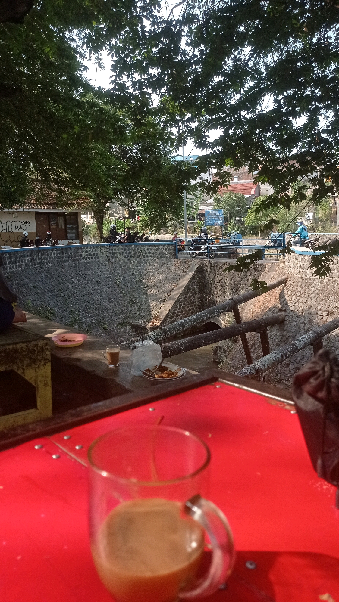 The image depicts a scene near a canal in Mulyoagung, Indonesia. In the foreground, a red table holds a clear glass mug filled with a light brown beverage, likely coffee or tea. Next to it, a small plate with what appears to be fried food rests on top of a plastic bag. A small pink plate with some food is also visible. The canal itself is lined with stone walls and has two large pipes running across it. Beyond the canal, a road is visible with several motorcycles and riders. Some buildings and trees line the far side of the road. The overall lighting suggests it is daytime, possibly late afternoon, with sunlight casting shadows.