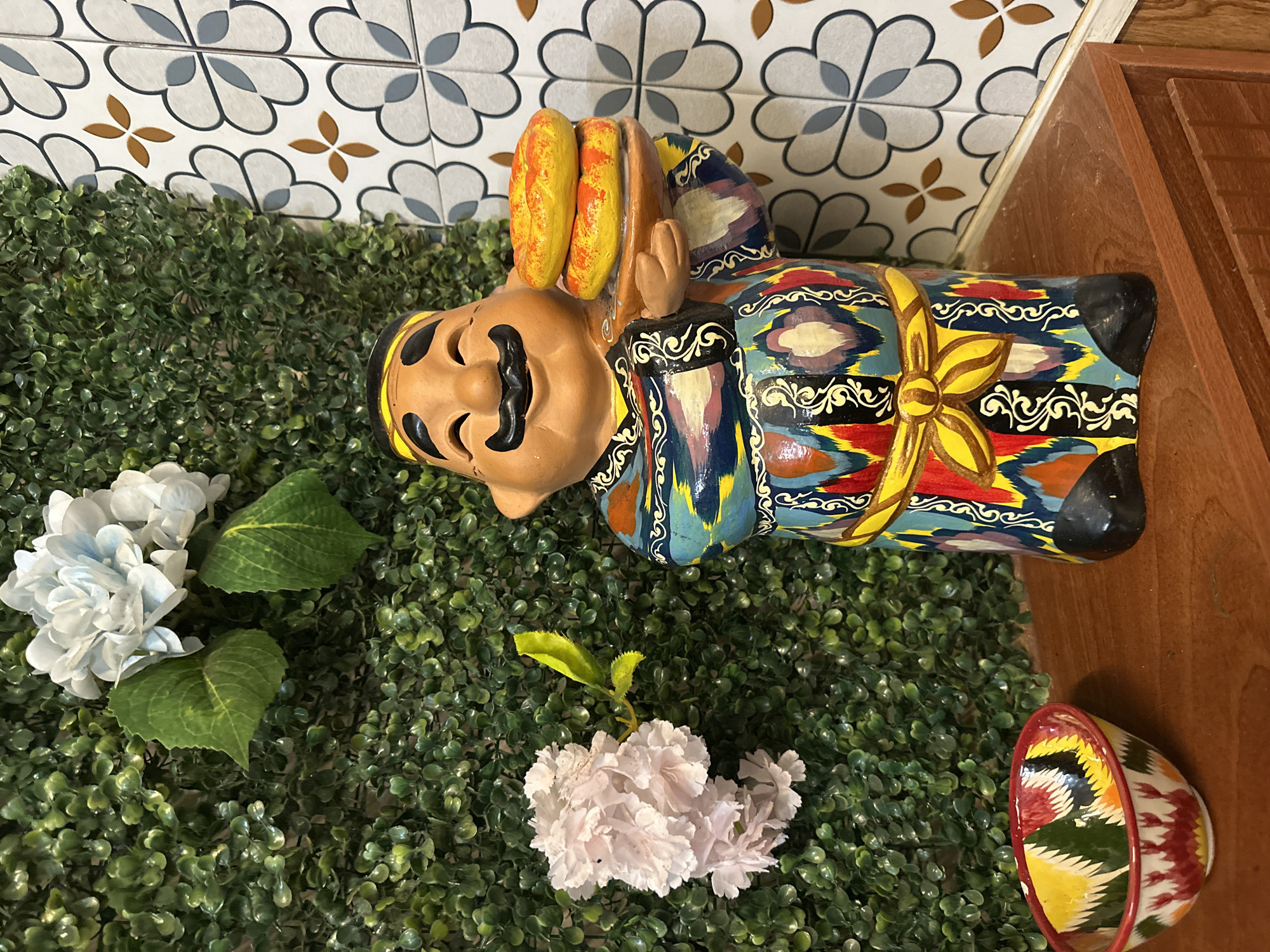 The image displays a vibrant decorative arrangement, presented as if rotated 90 degrees clockwise. At the center is a ceramic figurine of a smiling man with a prominent mustache, depicted lying on its side. He wears a richly patterned robe featuring bold geometric designs in shades of blue, yellow, red, black, and white, accented with a yellow sash or ribbon around his waist. The figurine carries two elongated, bread-like items, colored yellow and orange, over its left shoulder.
The figurine and artificial flowers rest on a surface covered with dense, artificial green foliage. To the left, a cluster of light blue and white artificial flowers with green leaves is visible. Another smaller cluster of light pink artificial blossoms with a single green stem appears near the bottom-center.
Behind the foliage, a wall is adorned with decorative white tiles featuring a repeating quatrefoil-like pattern in grey and brown. To the right, a wooden surface or piece of furniture is partially visible. In the bottom right corner, a small, colorful ceramic bowl with intricate geometric patterns in red, yellow, green, and black, and a red rim, is also present. The scene is indoors, brightly lit, and depicts no activity. There is no visible text.